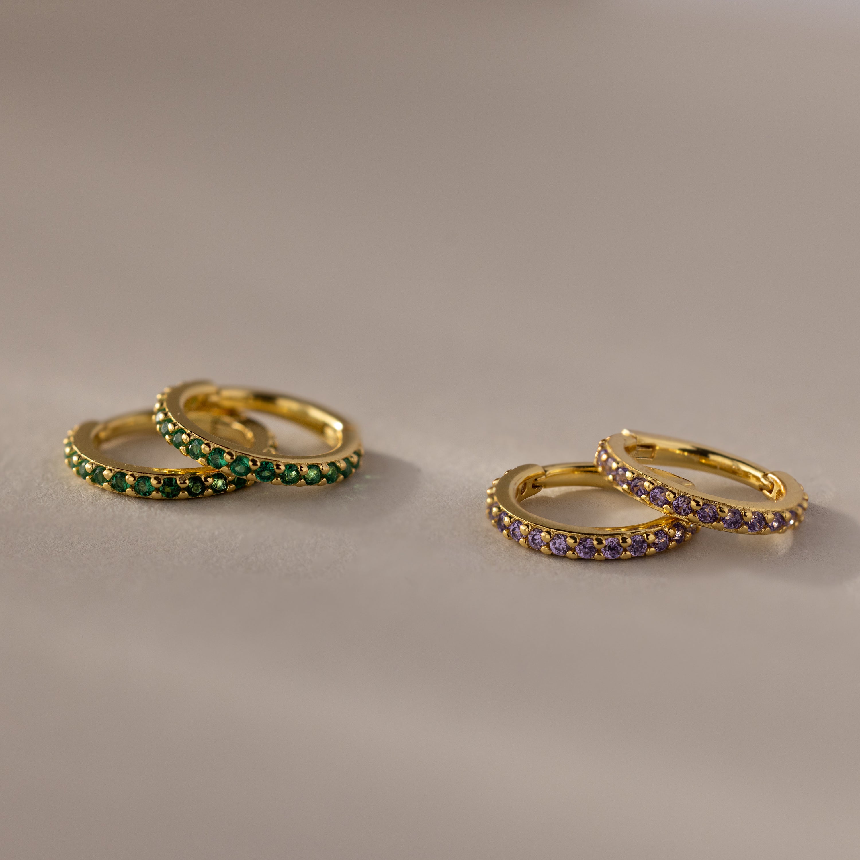 Two pairs of Birthstone Clicker Huggies—one with green stones, the other with purple—rest on a beige surface, making them perfect for everyday wear or as birthstone earrings.