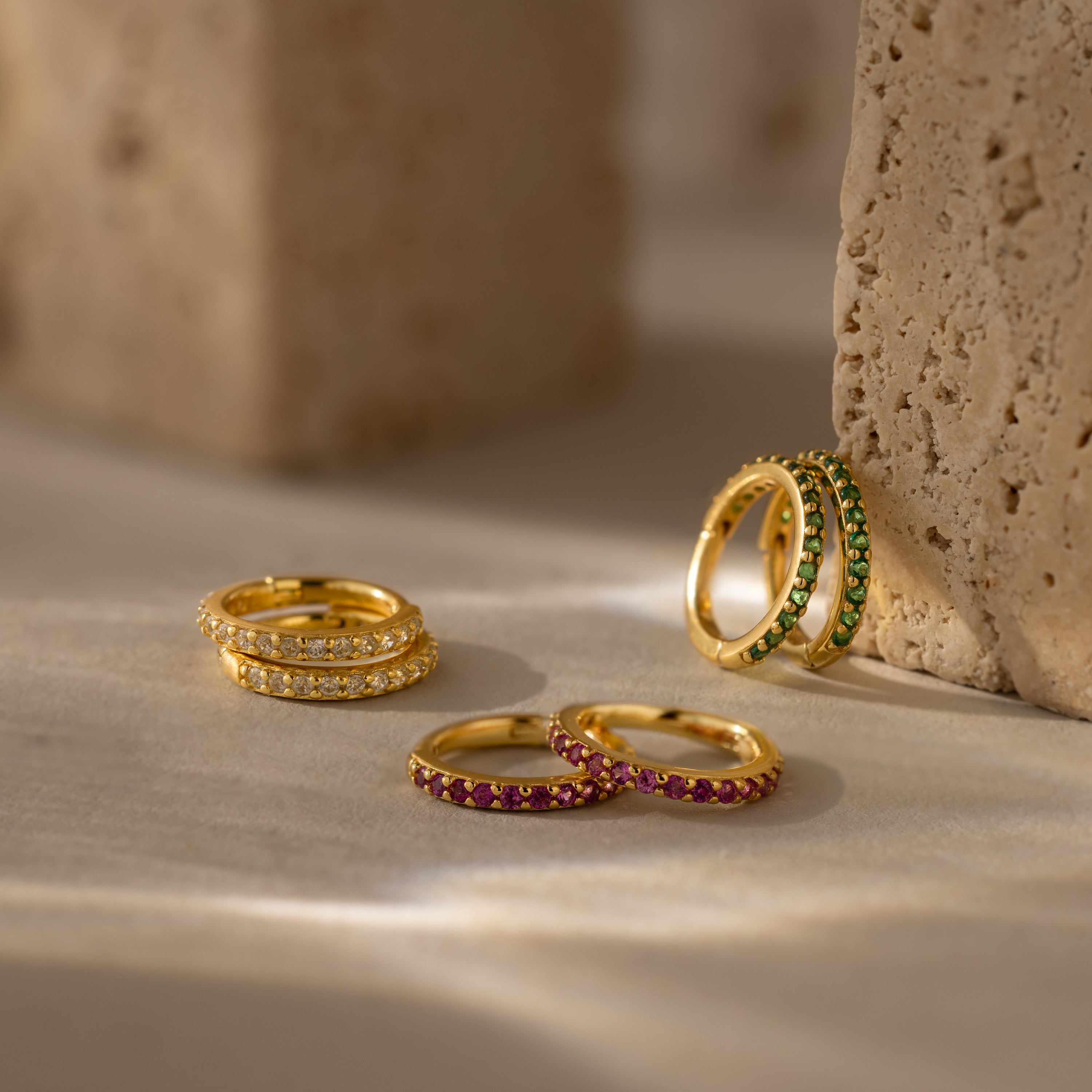 Four pairs of Diamond Clicker Huggies with white, green, and pink stones—perfect as birthstone earrings—are displayed on a neutral surface.