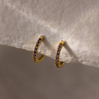 The Amethyst Clicker Huggies are gold hoop earrings with small purple amethyst stones, shown on a soft beige background—ideal as everyday huggies or as unique birthstone earrings for a personalized accessory.