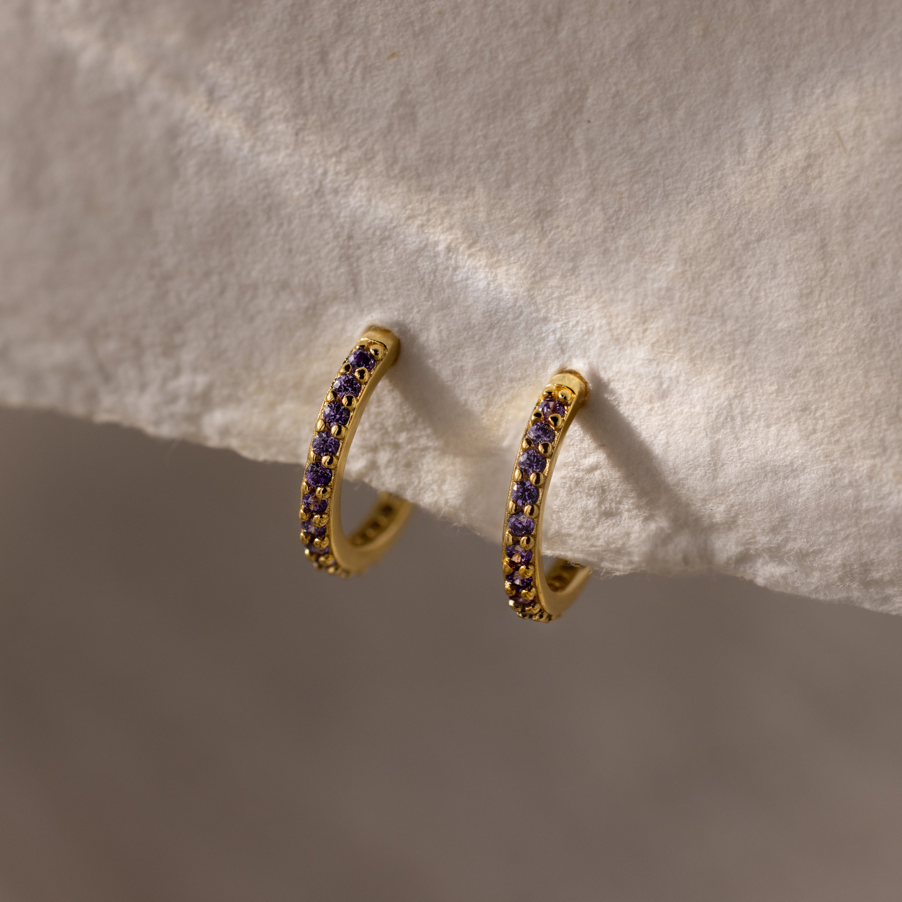 The Amethyst Clicker Huggies are gold hoop earrings with small purple amethyst stones, shown on a soft beige background—ideal as everyday huggies or as unique birthstone earrings for a personalized accessory.