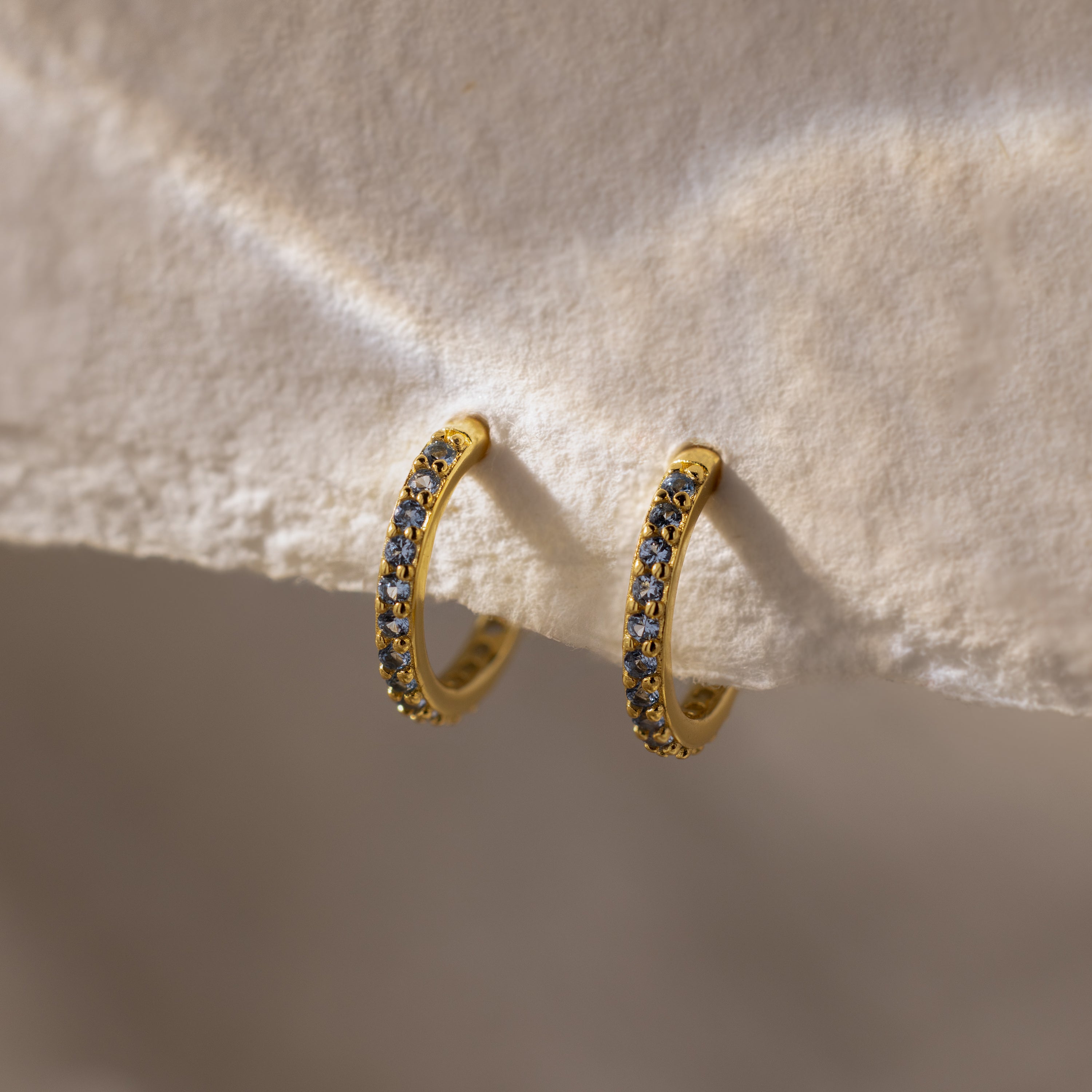 Aquamarine Clicker Huggies: gold hoop earrings adorned with small, sparkling aquamarine stones, perfect for everyday wear and shown on a textured, off-white surface.