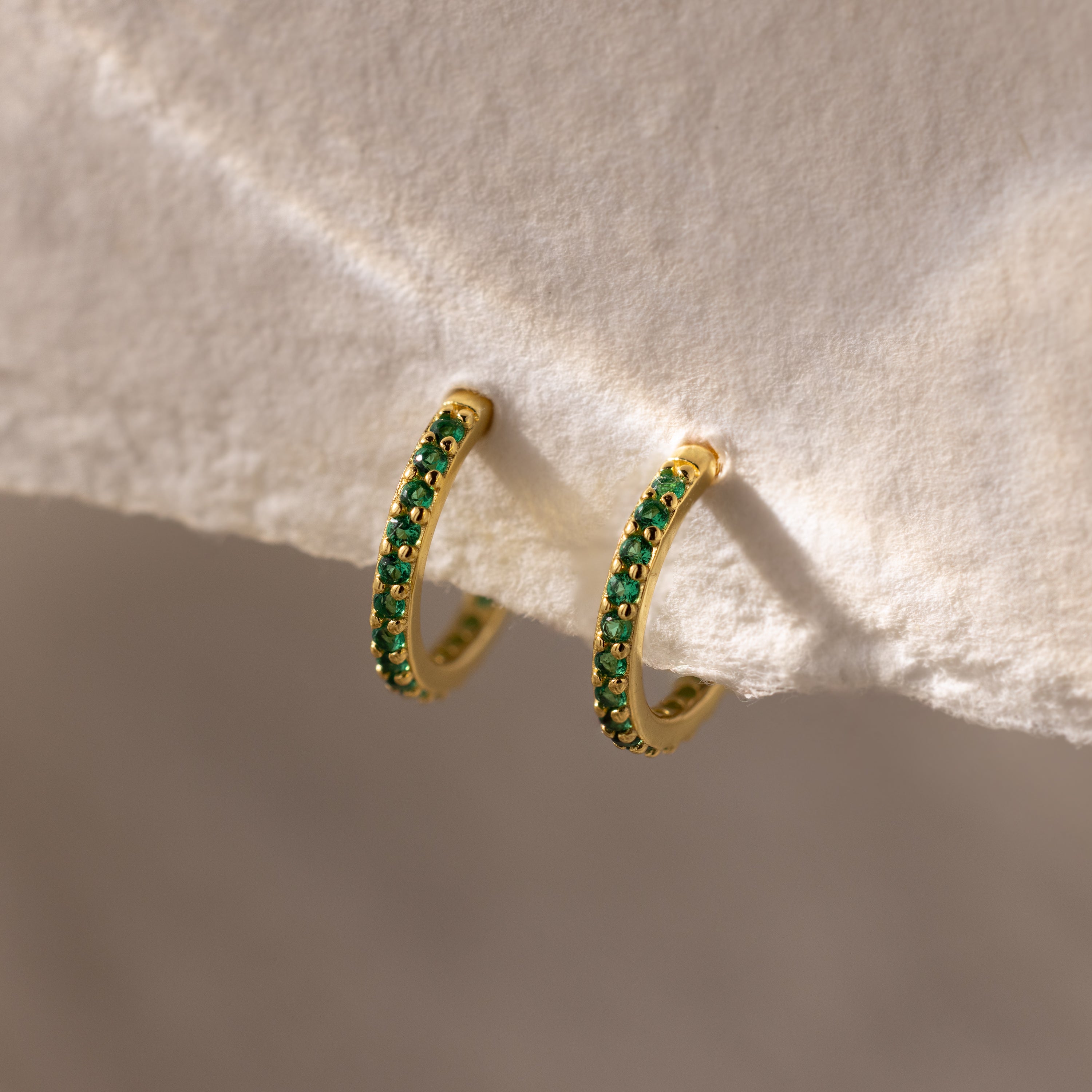 The Emerald Clicker Huggies are gold hoop earrings featuring small green gemstones, displayed on a textured off-white surface—perfect for everyday wear.