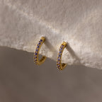 Alexandrite Clicker Huggies featuring gold hoops with small purple gemstones are showcased on a cream-colored textured fabric—ideal as everyday jewelry for a subtle, personal touch.