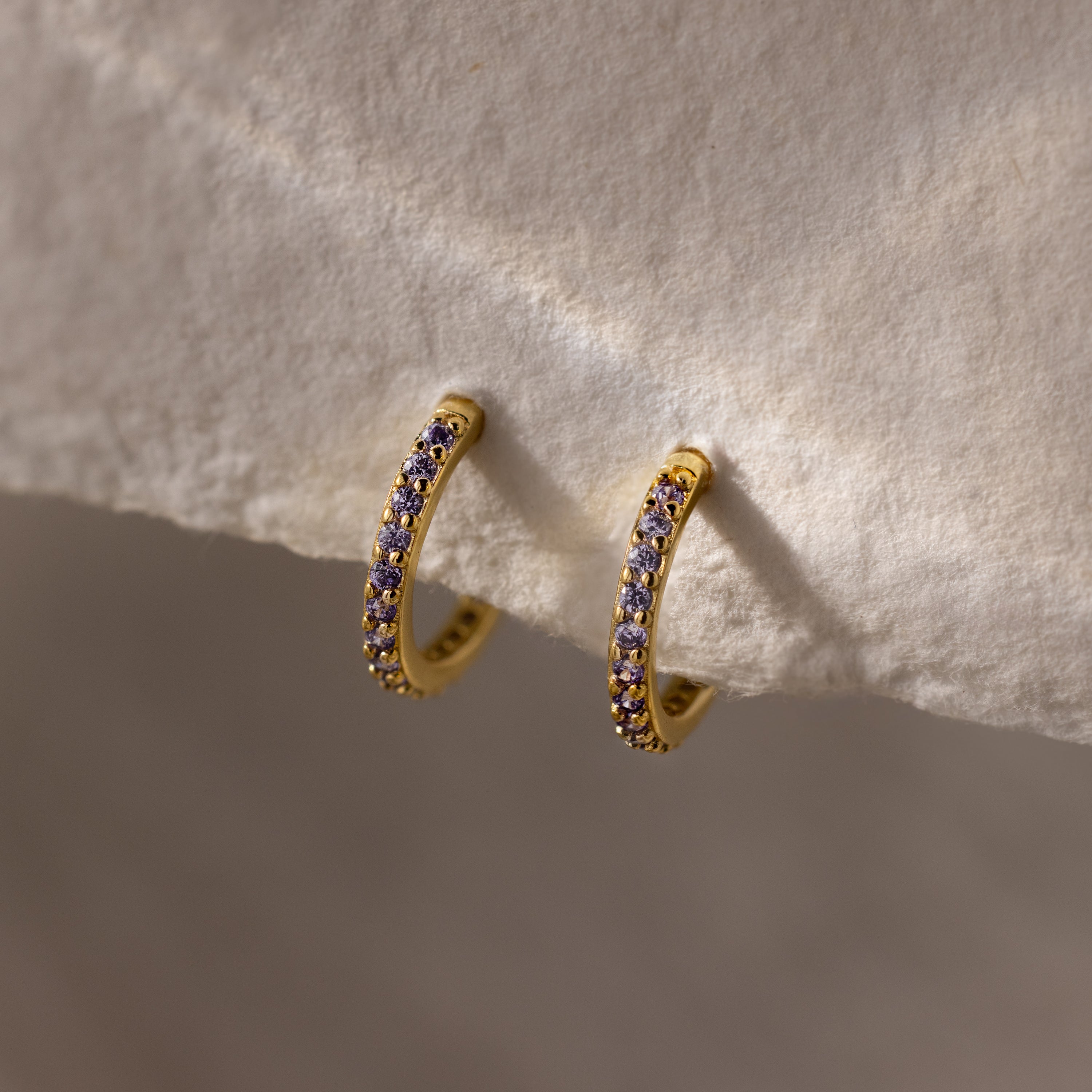 Alexandrite Clicker Huggies featuring gold hoops with small purple gemstones are showcased on a cream-colored textured fabric—ideal as everyday jewelry for a subtle, personal touch.