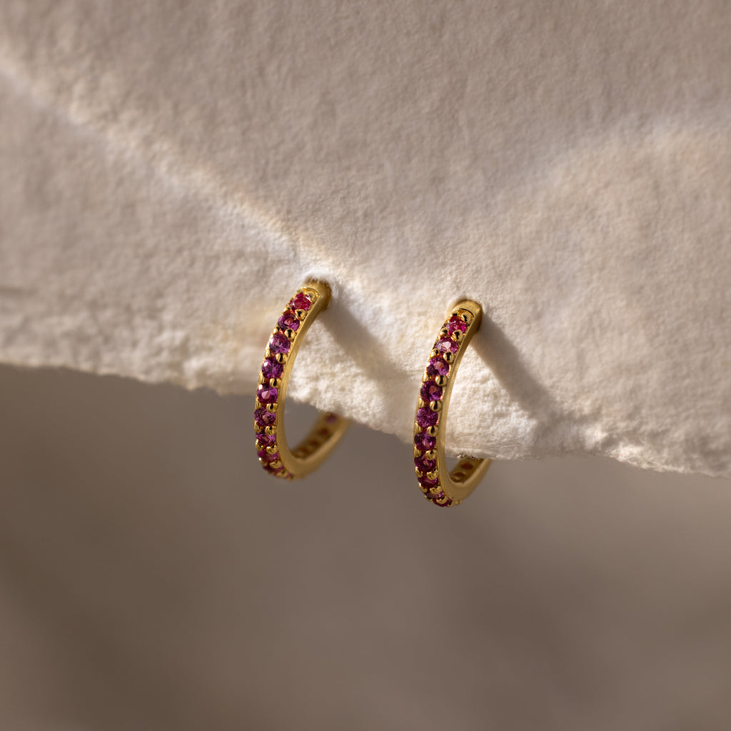 Ruby Clicker Huggies with small pink gemstones, shown on a textured beige surface—ideal for everyday wear or as thoughtful ruby birthstone earrings.