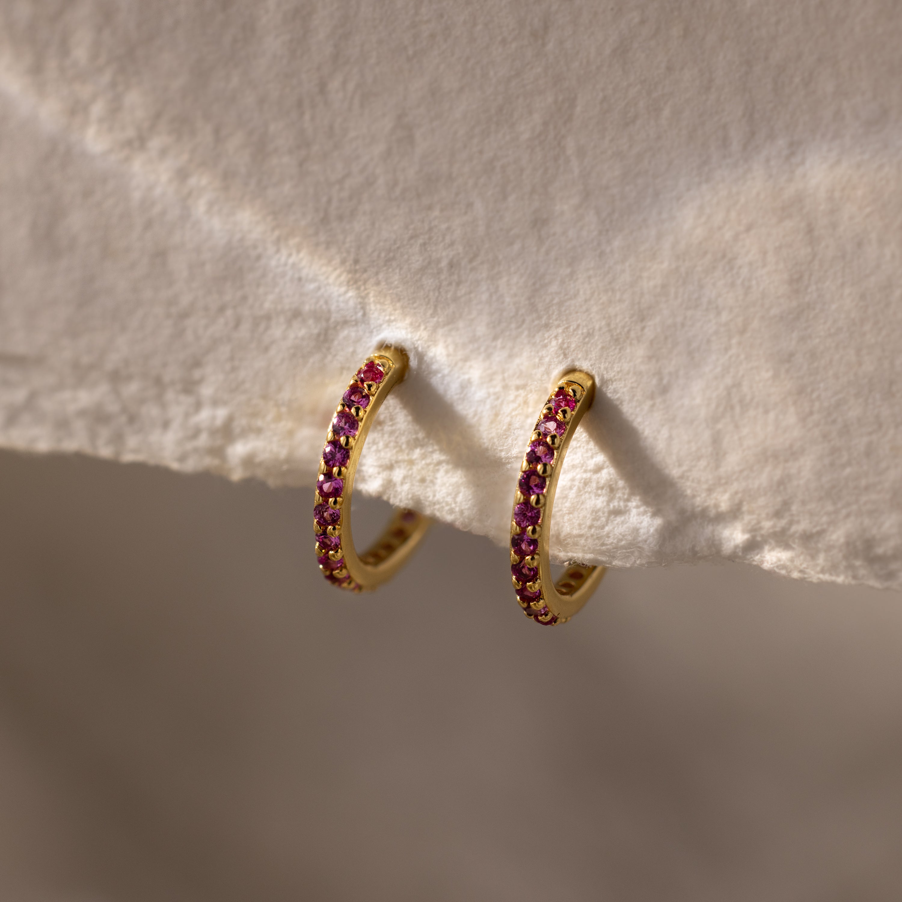 Ruby Clicker Huggies with small pink gemstones, shown on a textured beige surface—ideal for everyday wear or as thoughtful ruby birthstone earrings.
