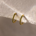 Peridot Clicker Huggies—gold hoop earrings with small green gemstones, shown on a textured white background—make perfect birthstone earrings to add a pop of color to any everyday outfit.