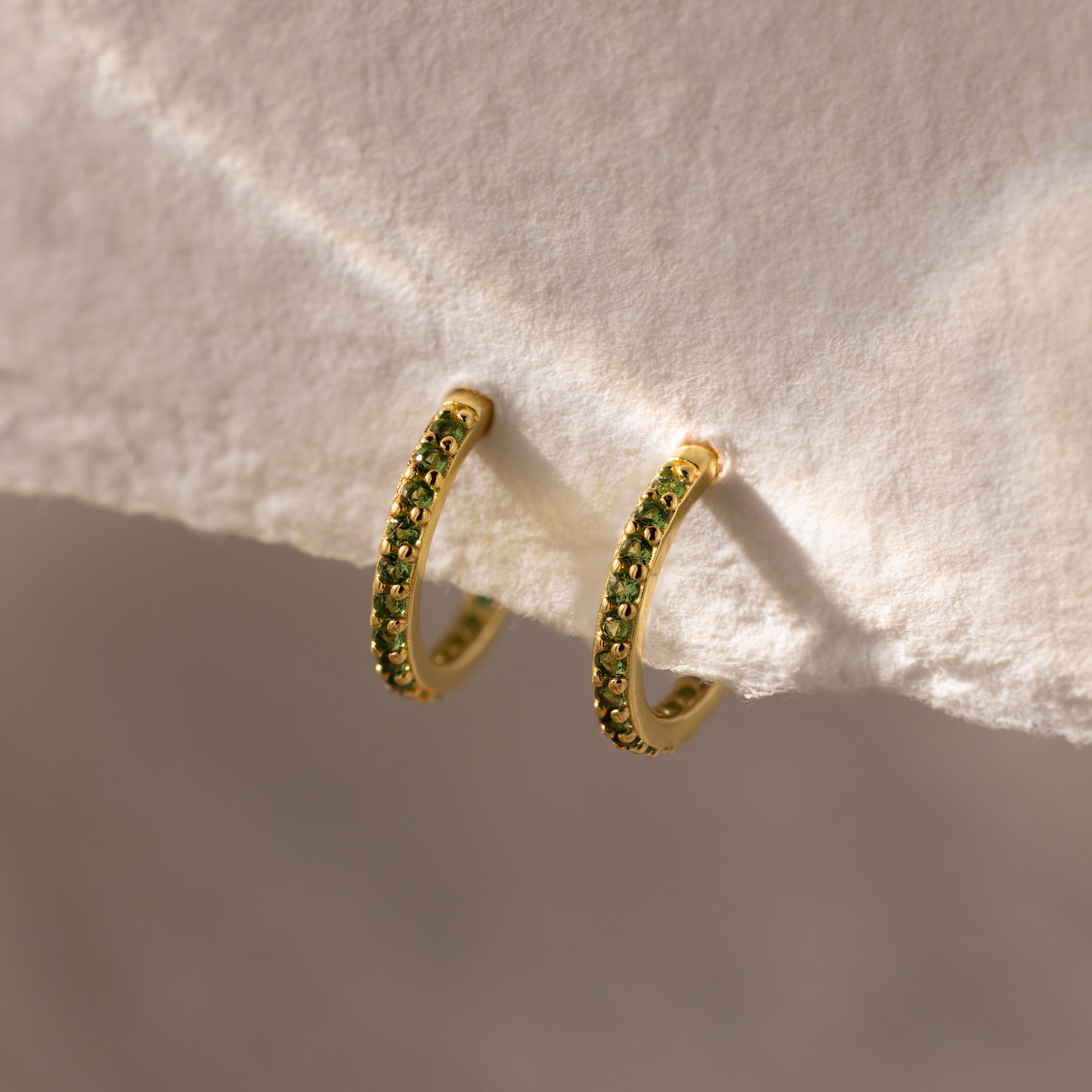 Peridot Clicker Huggies—gold hoop earrings with small green gemstones, shown on a textured white background—make perfect birthstone earrings to add a pop of color to any everyday outfit.