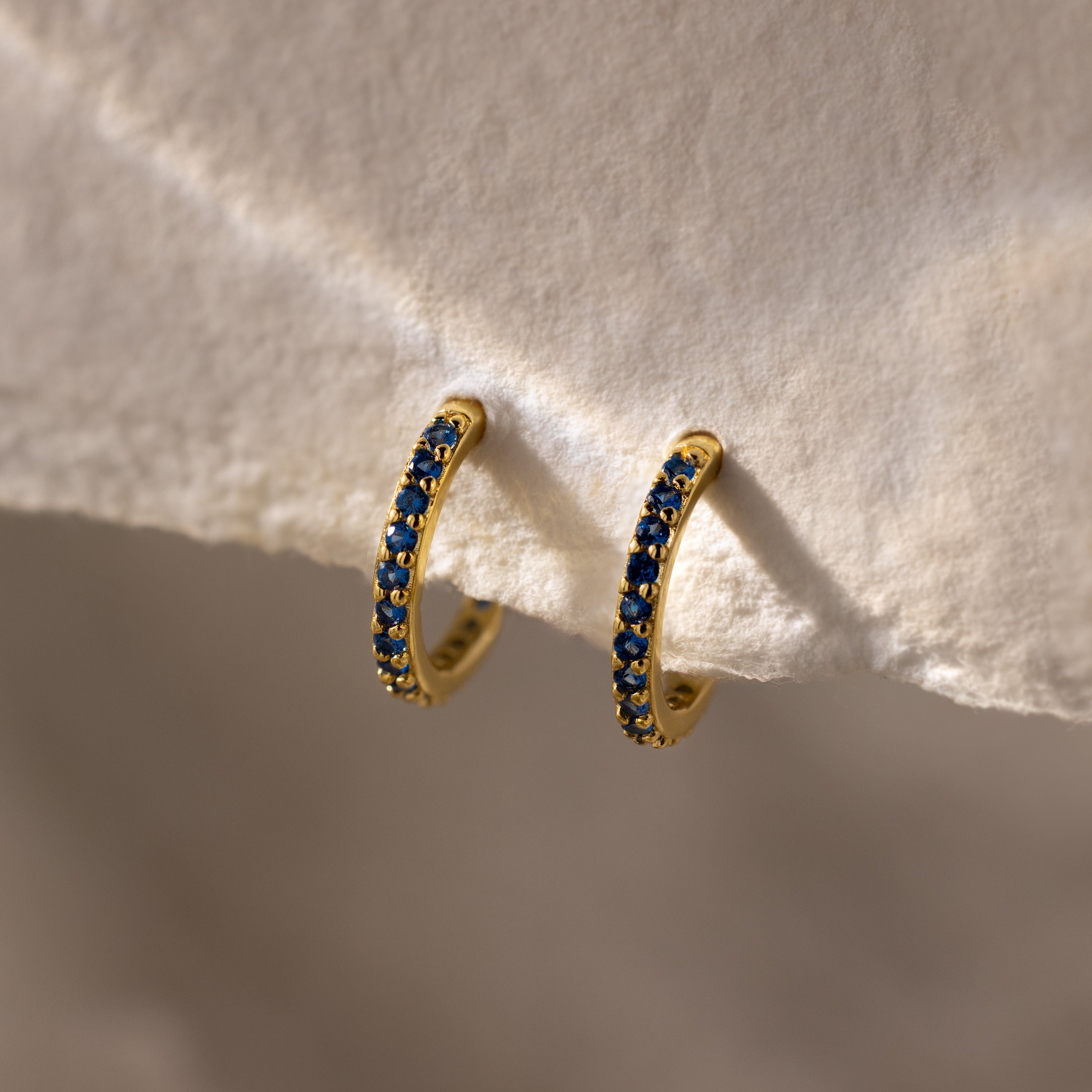 The Sapphire Clicker Huggies are gold hoop earrings featuring a row of small blue gemstones, perfect for everyday wear and shown on a textured cream surface.