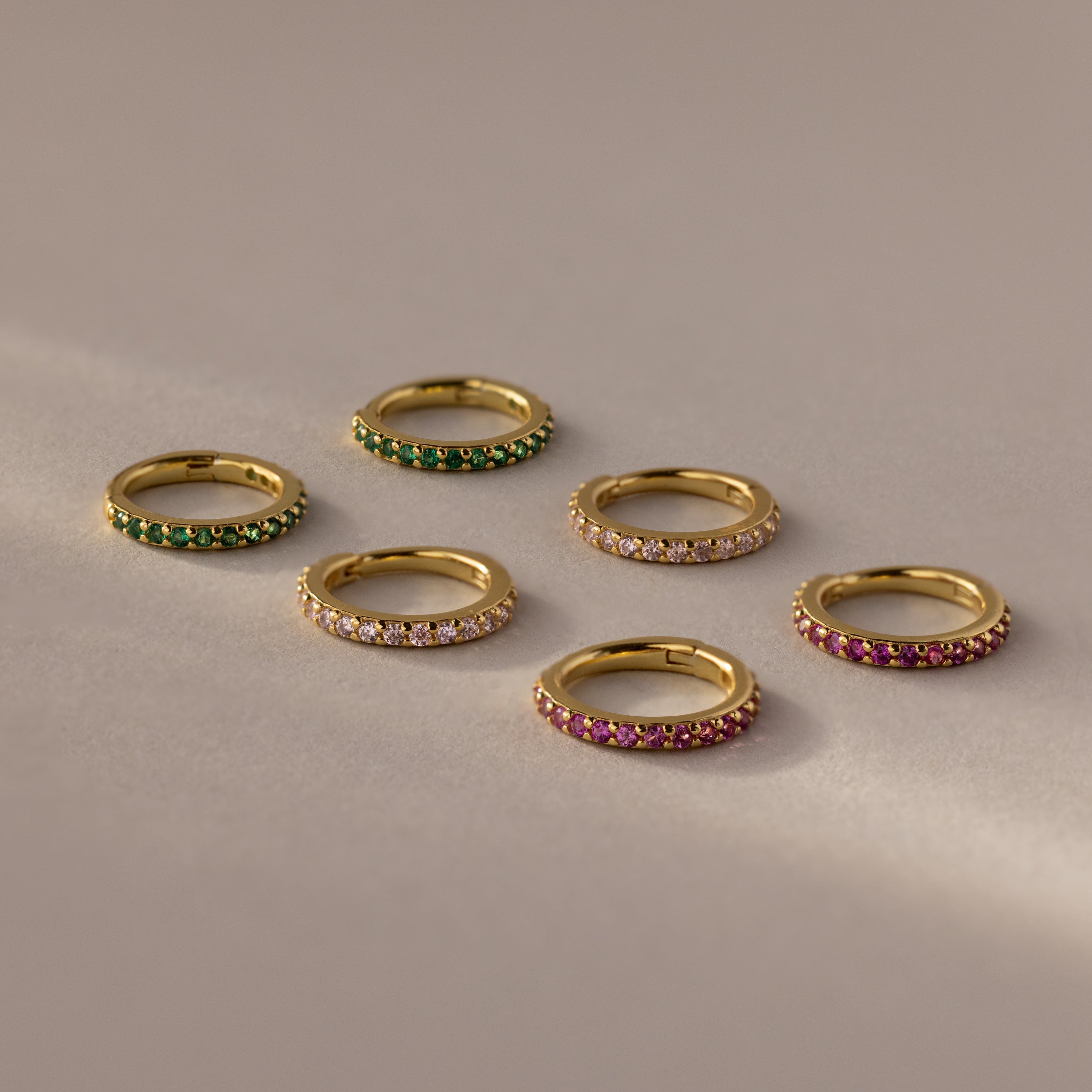 Six Birthstone Clicker Huggies with green, clear, and pink gemstones arranged on a light beige surface—perfect for stacking with your favorite everyday jewelry or pairing with birthstone earrings.