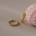 Two Birthstone Clicker Huggies featuring small pink gemstones—one flat, one leaning on a textured pink seashell—make the perfect everyday jewelry.