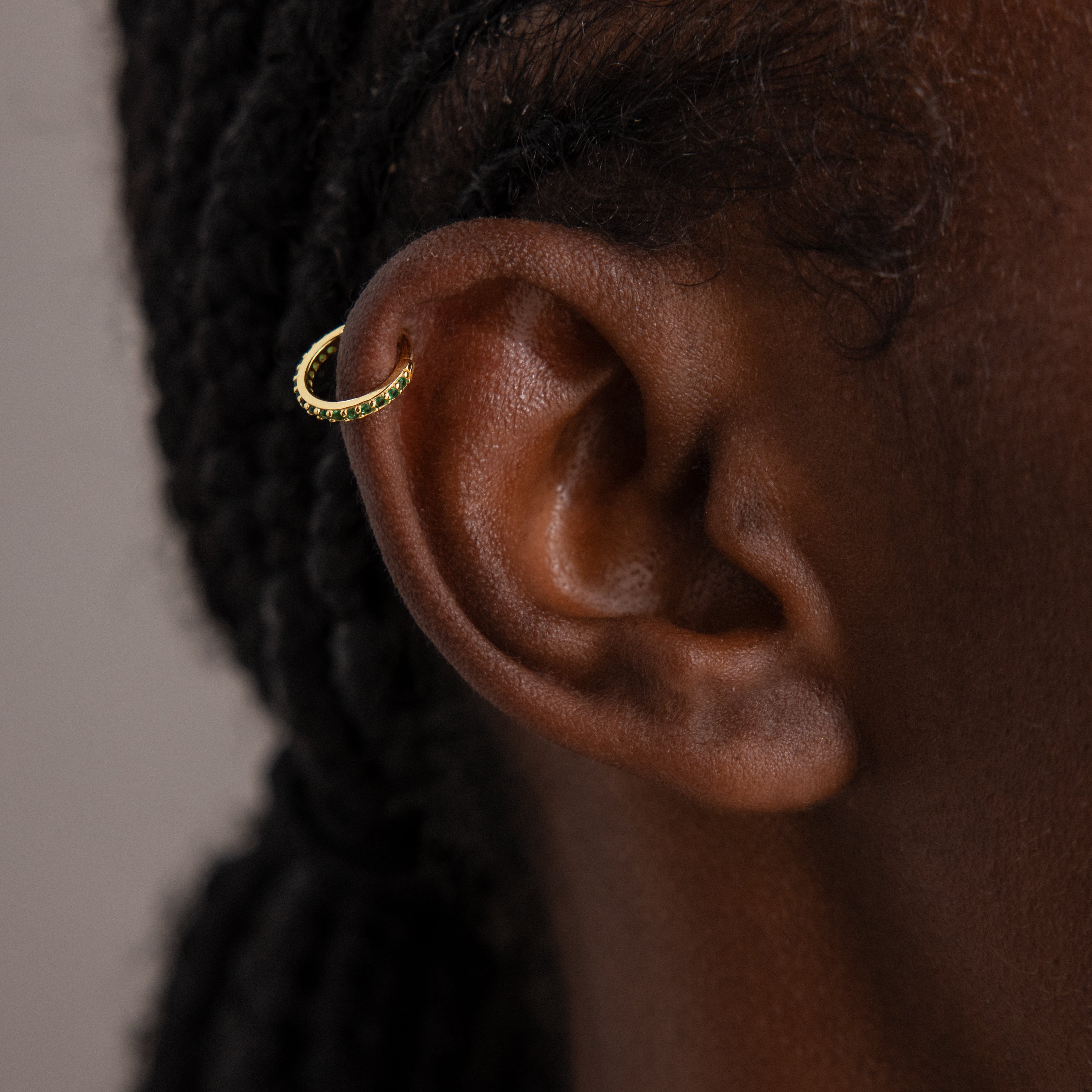 Close-up of an ear wearing Birthstone Clicker Huggies, elegant gold hoop earrings ideal for daily wear; braided hair appears in the background.