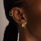 Close-up of an ear adorned with multiple gold earrings, featuring the Birthstone Clicker Huggies with a green gem—ideal for everyday wear—with braided hair visible in the background.