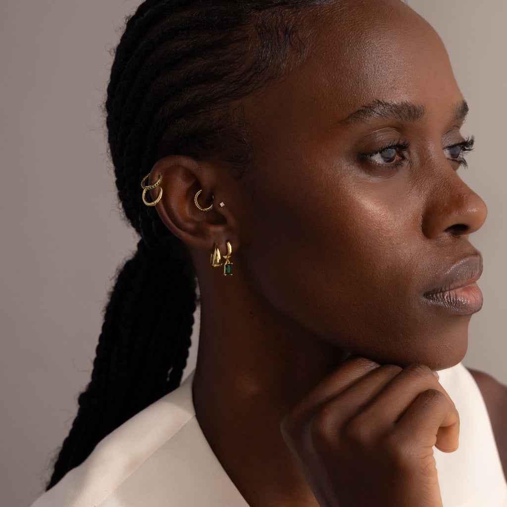 A woman with braided hair, wearing multiple gold earrings including elegant Birthstone Clicker Huggies, rests her chin on her hand and looks thoughtful.