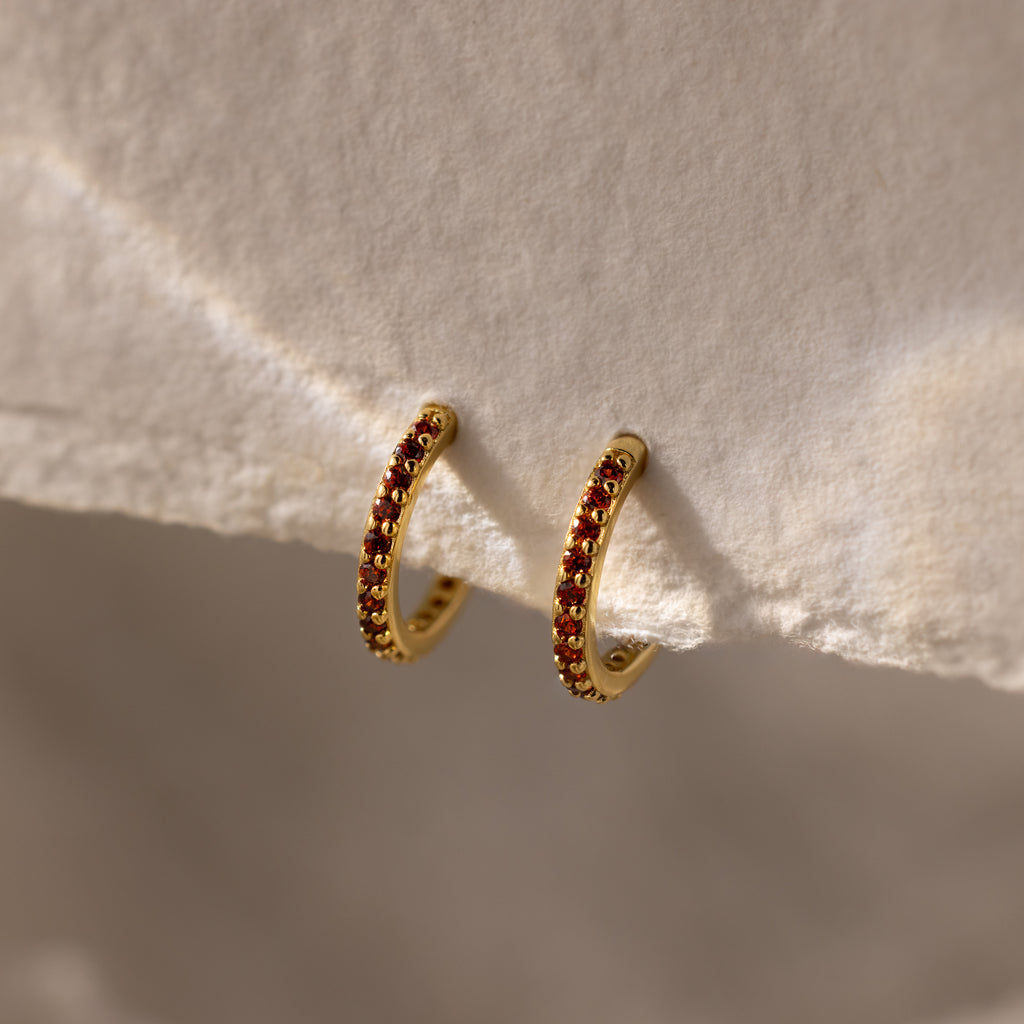 The Garnet Clicker Huggies are gold hoop earrings featuring small red gemstones, shown on a textured cream surface—perfect as birthstone earrings and ideal for everyday wear.