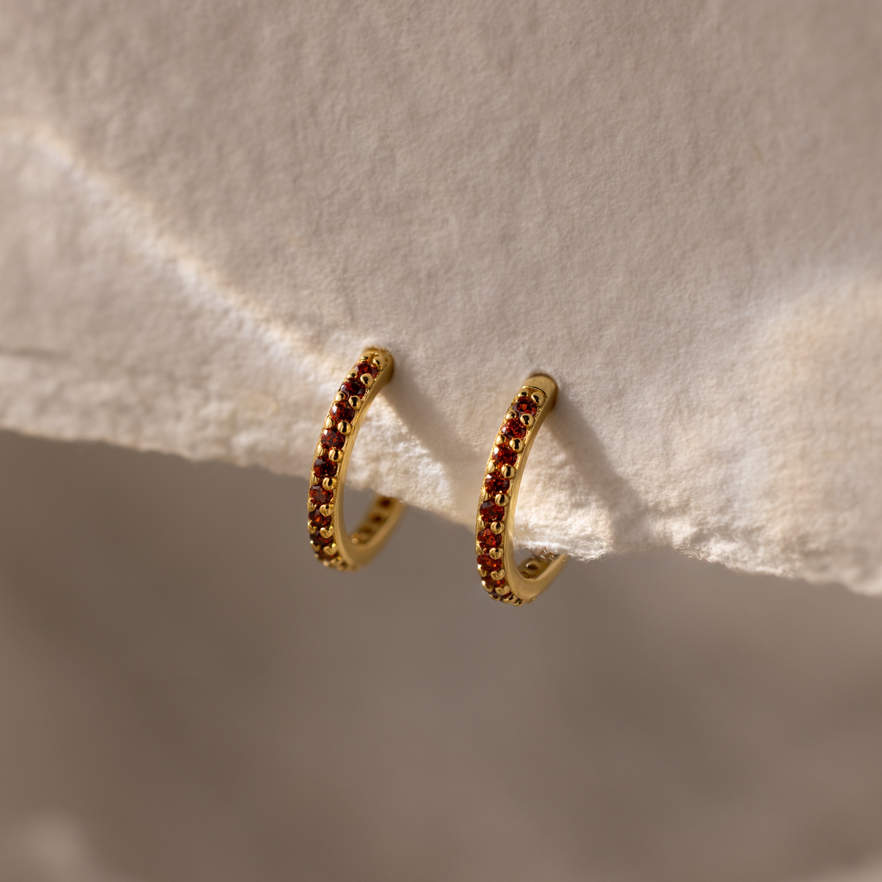 The Garnet Clicker Huggies are gold hoop earrings featuring small red gemstones, shown on a textured cream surface—perfect as birthstone earrings and ideal for everyday wear.