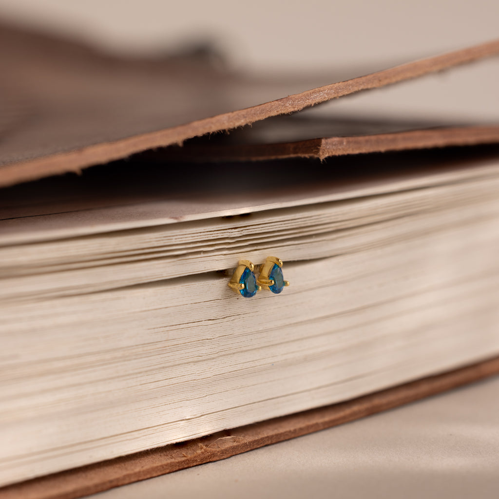 The Teardrop Blue Zircon Flatback Studs, in elegant blue and gold, rest on a closed book, bringing a touch of classic style to any look.