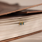 The Teardrop Blue Zircon Flatback Studs, in elegant blue and gold, rest on a closed book, bringing a touch of classic style to any look.