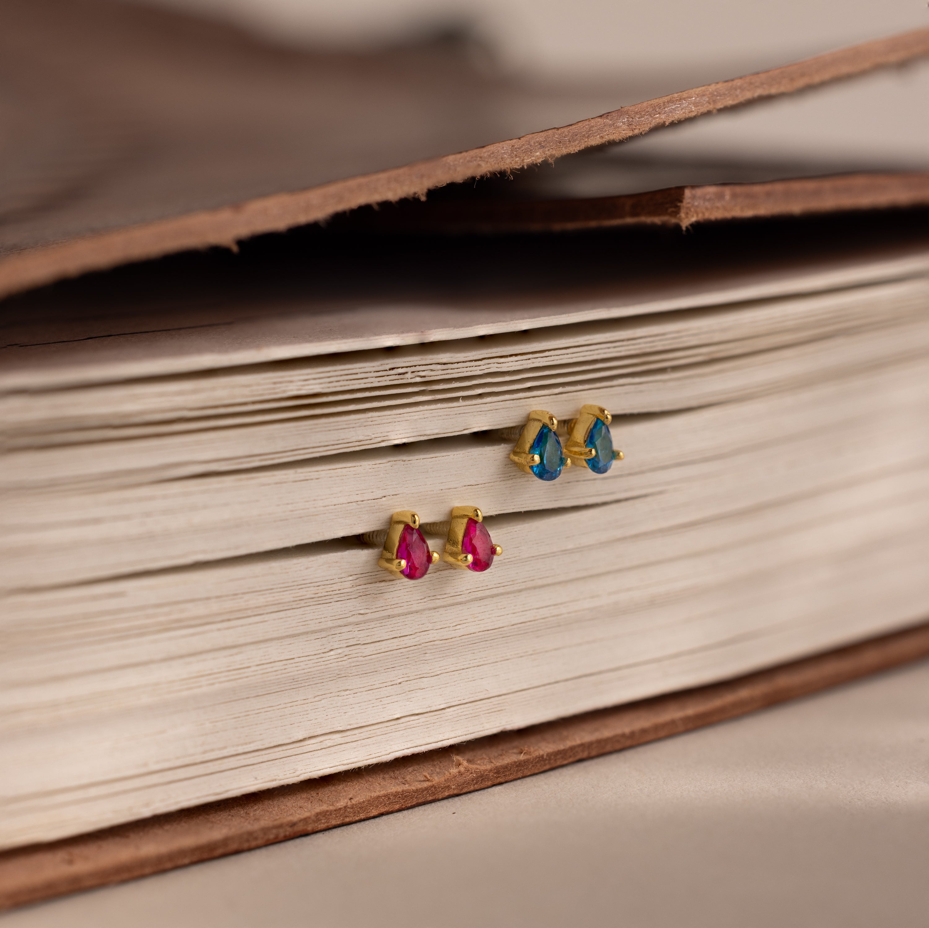 Teardrop Blue Zircon Flatback Studs with gold finish and sparkling blue gems bring a touch of elegant birthstone jewelry as they rest delicately on the pages of an open book.