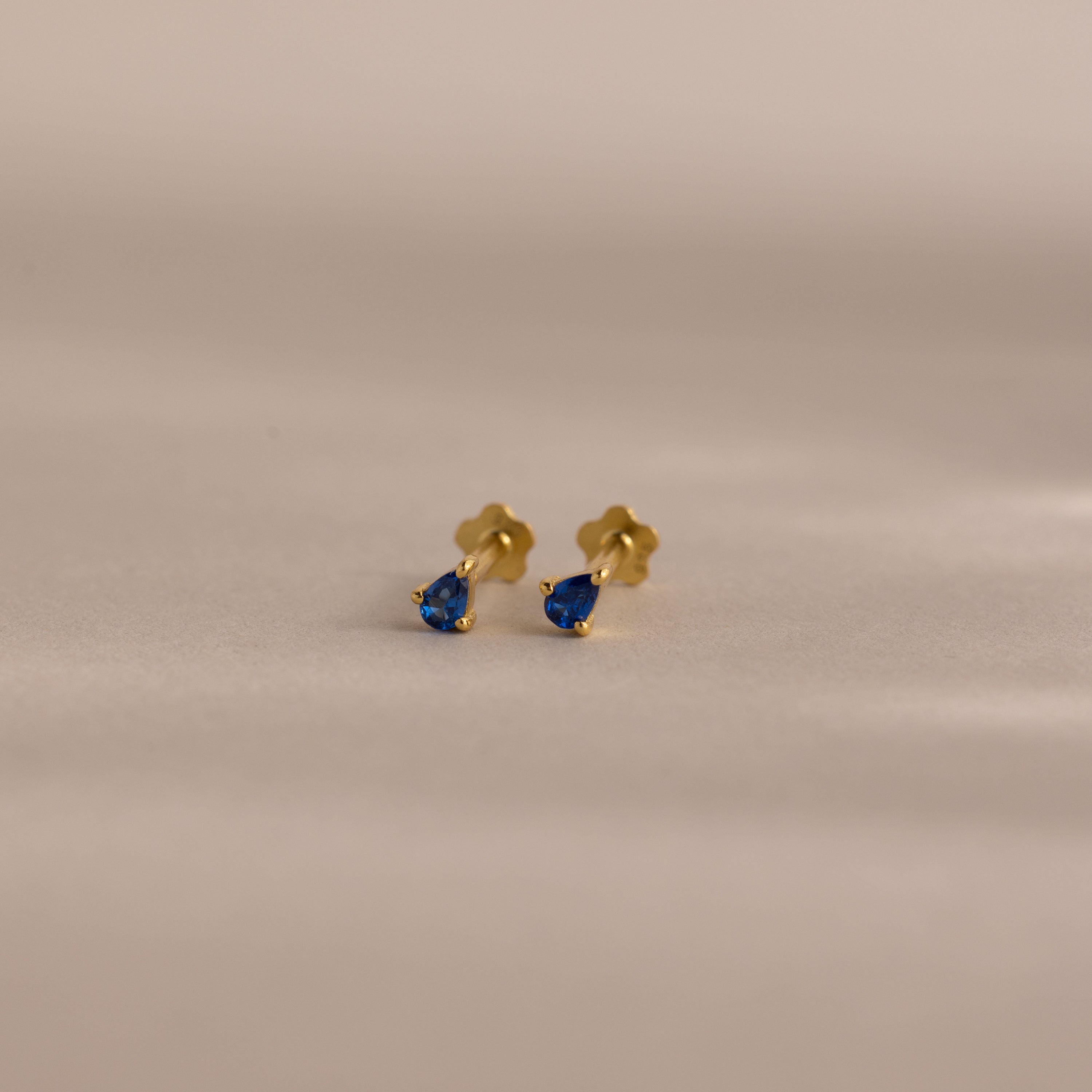 Teardrop Sapphire Flatback Studs featuring gold and small blue teardrop gems—a stunning piece of birthstone jewelry, showcased on a neutral background.
