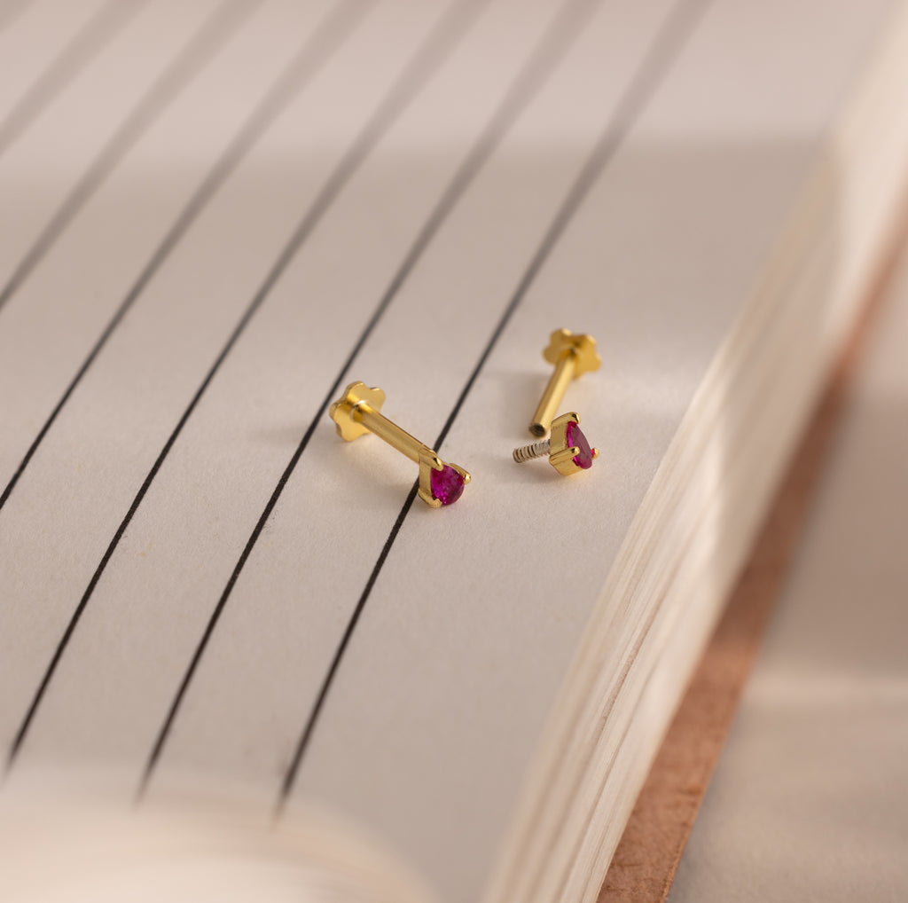 A pair of Teardrop Birthstone Flatback Studs with small red stones is displayed on the open pages of a lined notebook.
