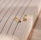 A pair of Teardrop Birthstone Flatback Studs with small red stones is displayed on the open pages of a lined notebook.