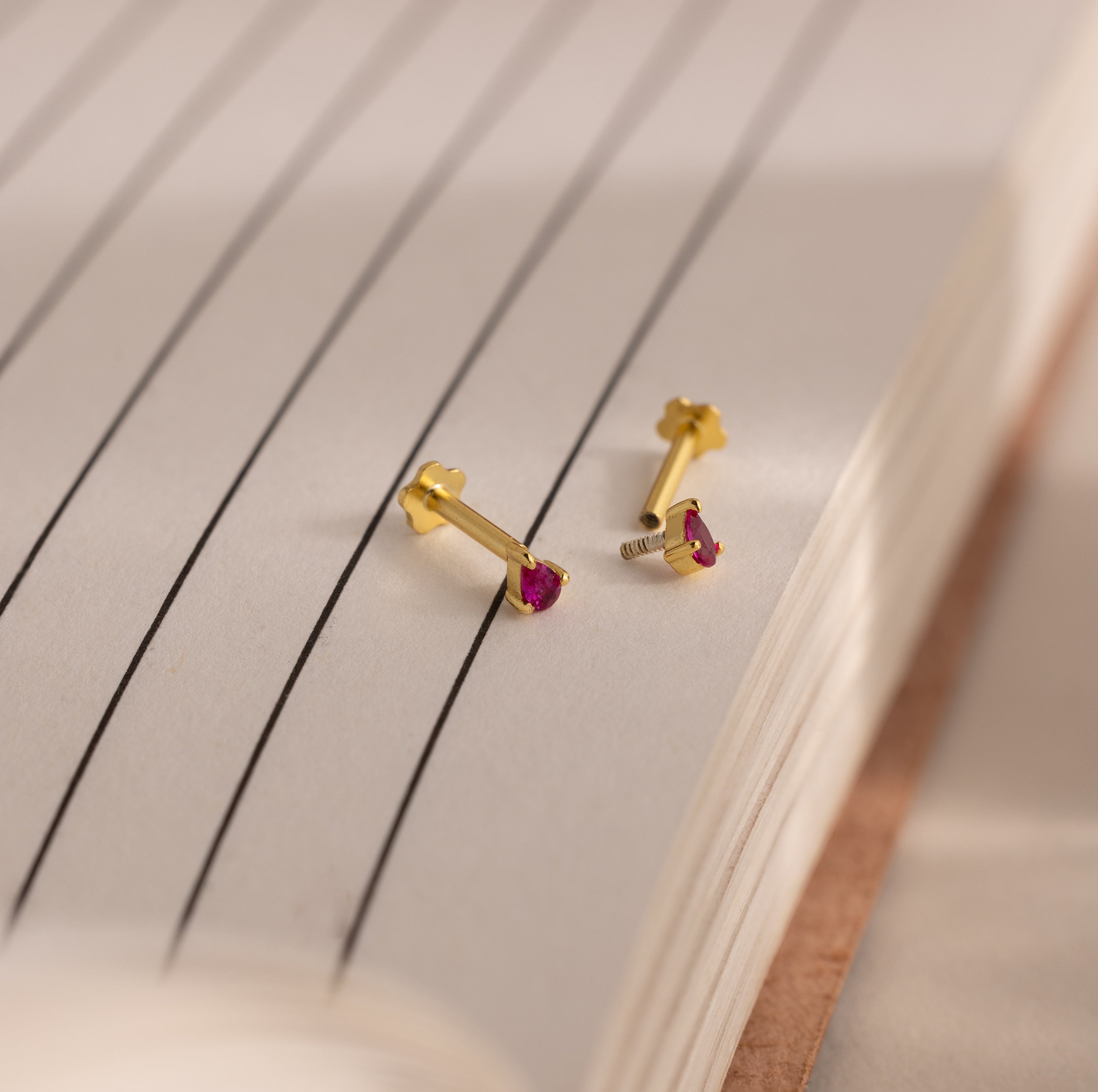 A pair of Teardrop Birthstone Flatback Studs with small red stones is displayed on the open pages of a lined notebook.