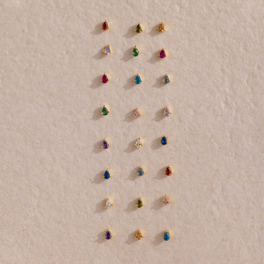 Teardrop Sapphire Flatback Studs are displayed in neat rows on a textured beige surface, showcasing a charming array of colorful birthstone jewelry.