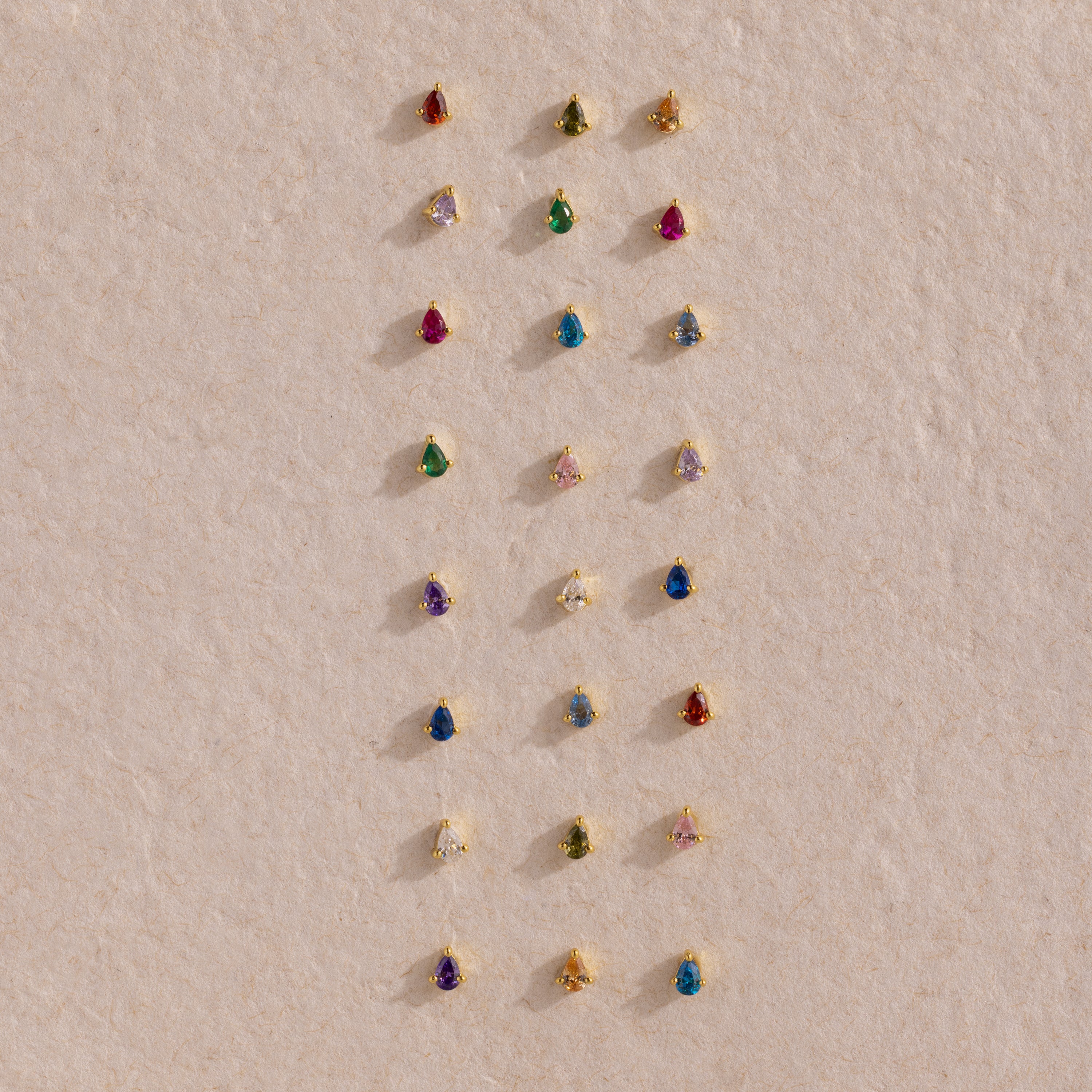 Teardrop Sapphire Flatback Studs are displayed in neat rows on a textured beige surface, showcasing a charming array of colorful birthstone jewelry.
