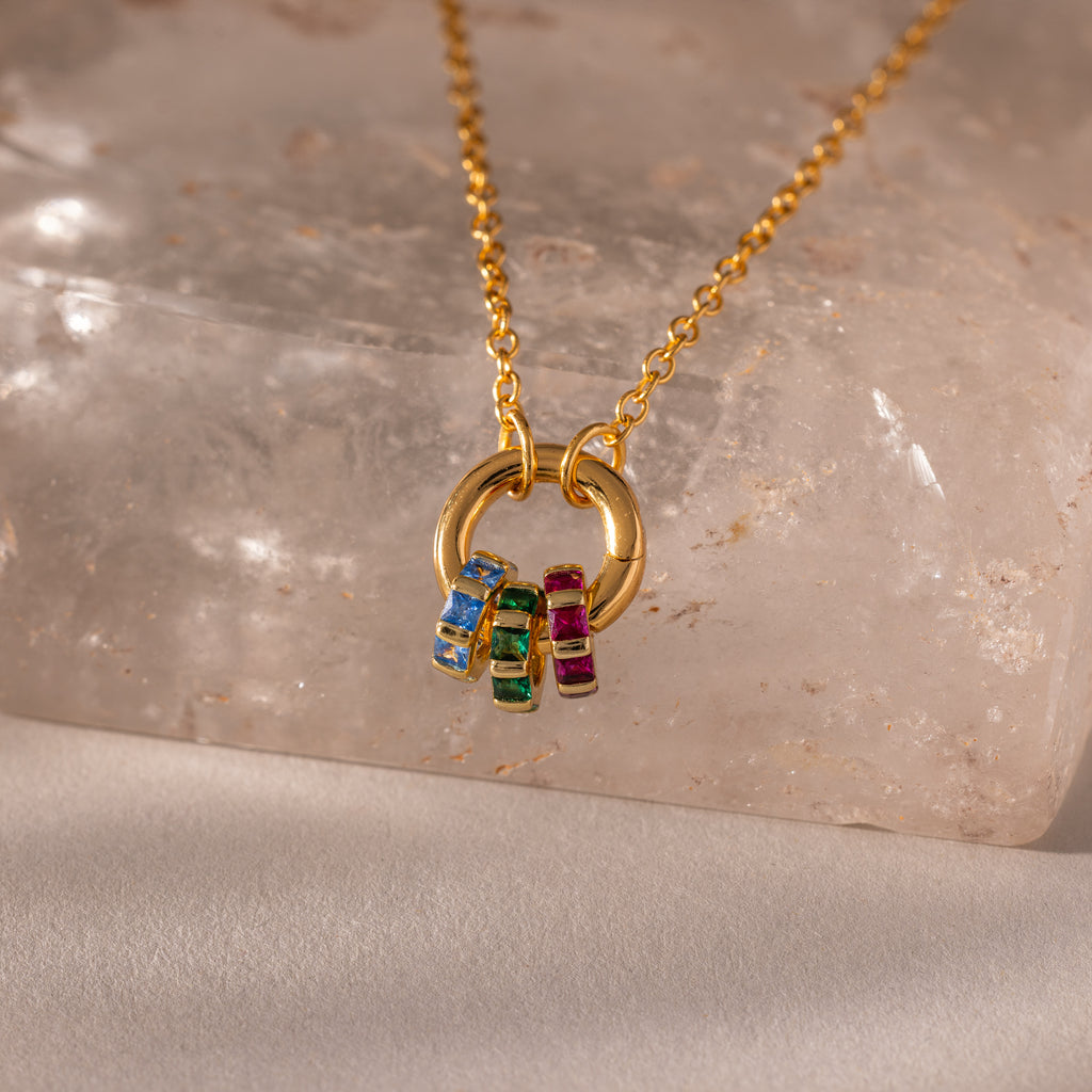 The Infinity Princess Birthstone Charm, a gold necklace adorned with colorful gemstone rings, shines beautifully on a light, textured surface.