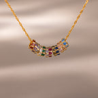 The Infinity Princess Birthstone Charm is a gold chain necklace featuring colorful, princess-cut stones in gemstone-filled rings, shown against a neutral background.