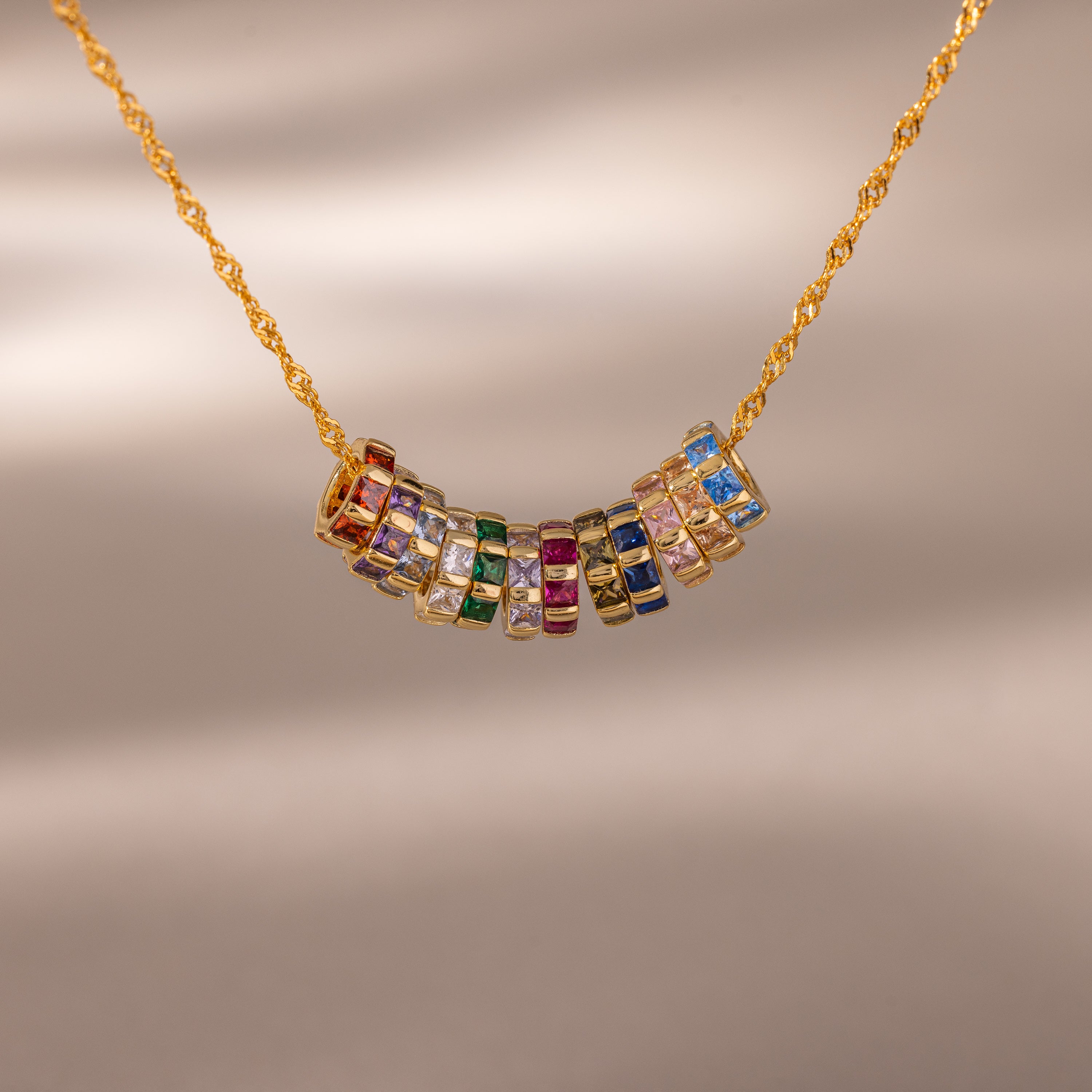 The Infinity Princess Birthstone Charm is a gold chain necklace featuring colorful, princess-cut stones in gemstone-filled rings, shown against a neutral background.