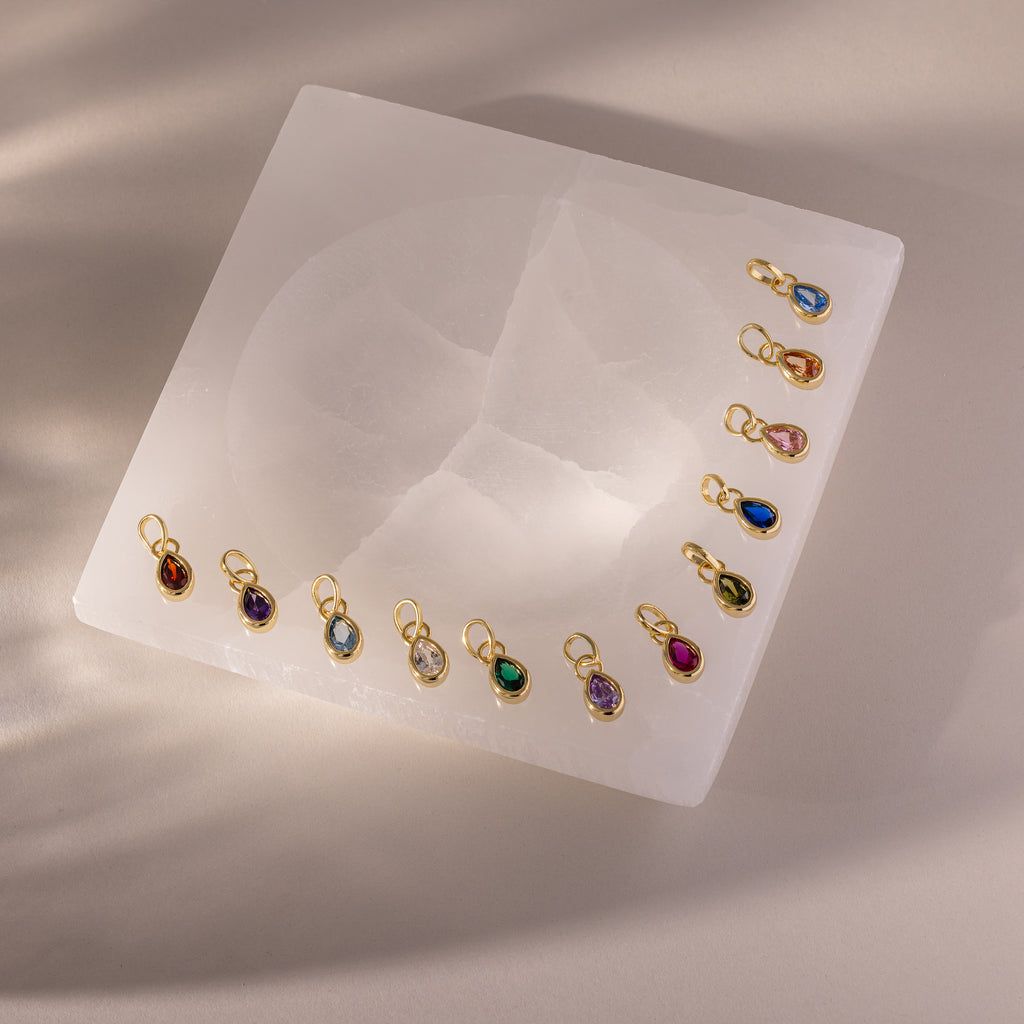 Twelve Teardrop Blue Zircon Charms, each set in a gold bezel with gold loops, are arranged on a square white stone surface.