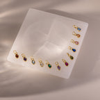 Twelve Teardrop Blue Zircon Charms, each set in a gold bezel with gold loops, are arranged on a square white stone surface.