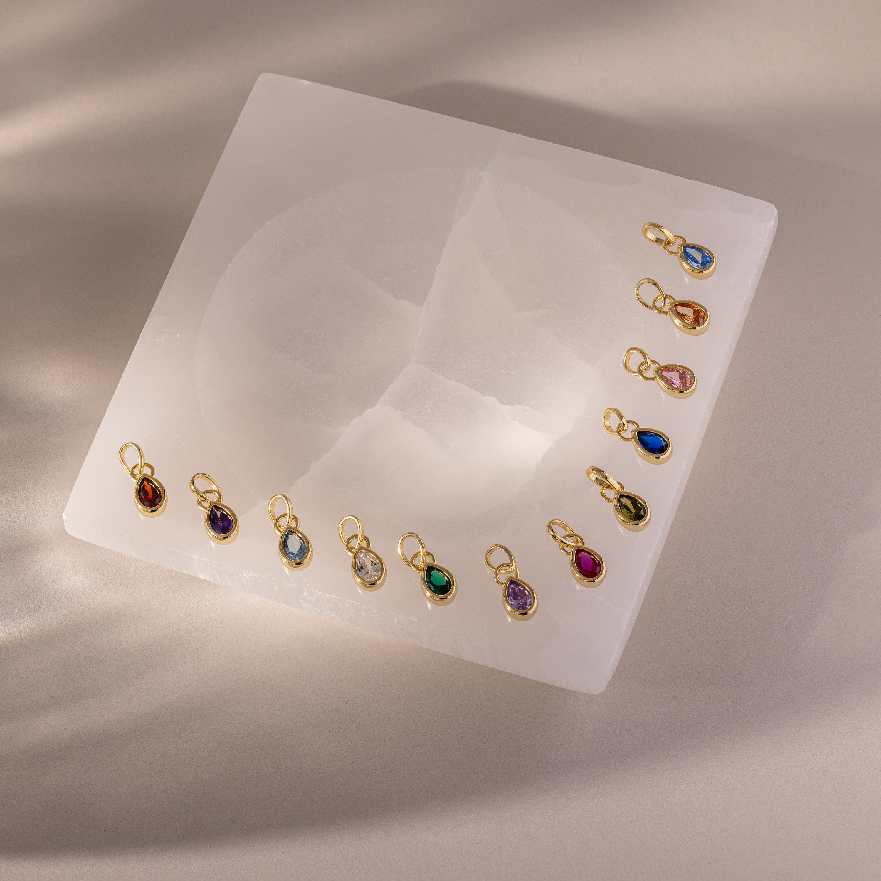 Twelve Teardrop Blue Zircon Charms, each set in a gold bezel with gold loops, are arranged on a square white stone surface.