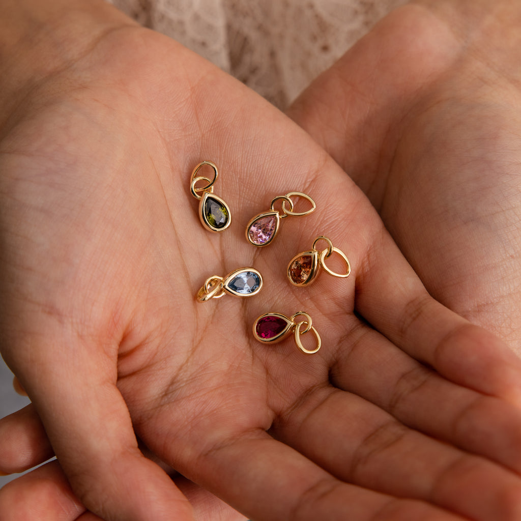 A hand holds six Teardrop Blue Zircon Charms, each featuring a vibrant gemstone in a gold bezel setting—ideal for creating personalized teardrop birthstone jewelry.