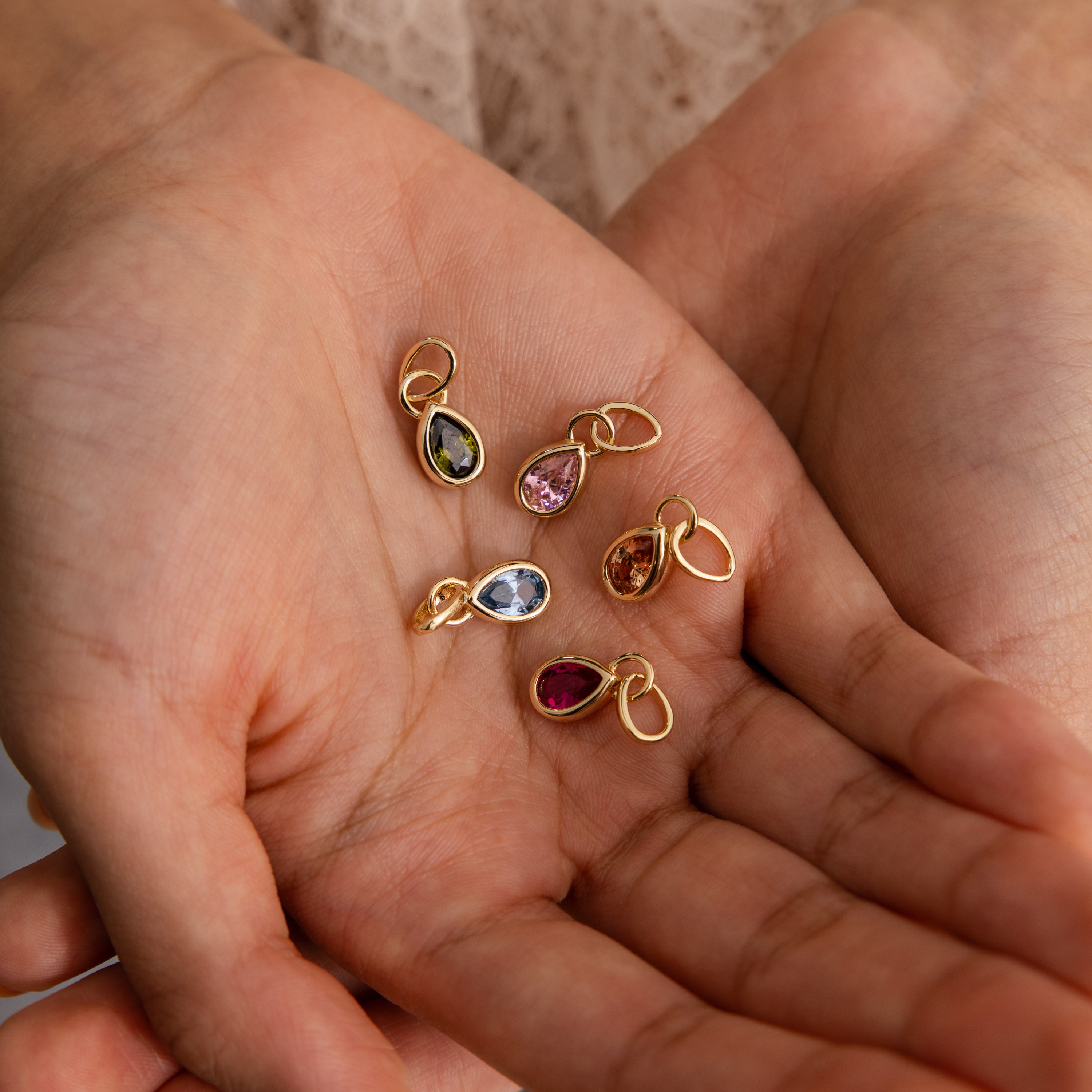 A hand holds six Teardrop Blue Zircon Charms, each featuring a vibrant gemstone in a gold bezel setting—ideal for creating personalized teardrop birthstone jewelry.