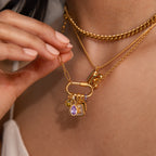 A woman's hand holds gold charm necklaces with gemstone pendants, including the Teardrop Birthstone Charm, a purple gem, letter charms, and a lock in a gold bezel.