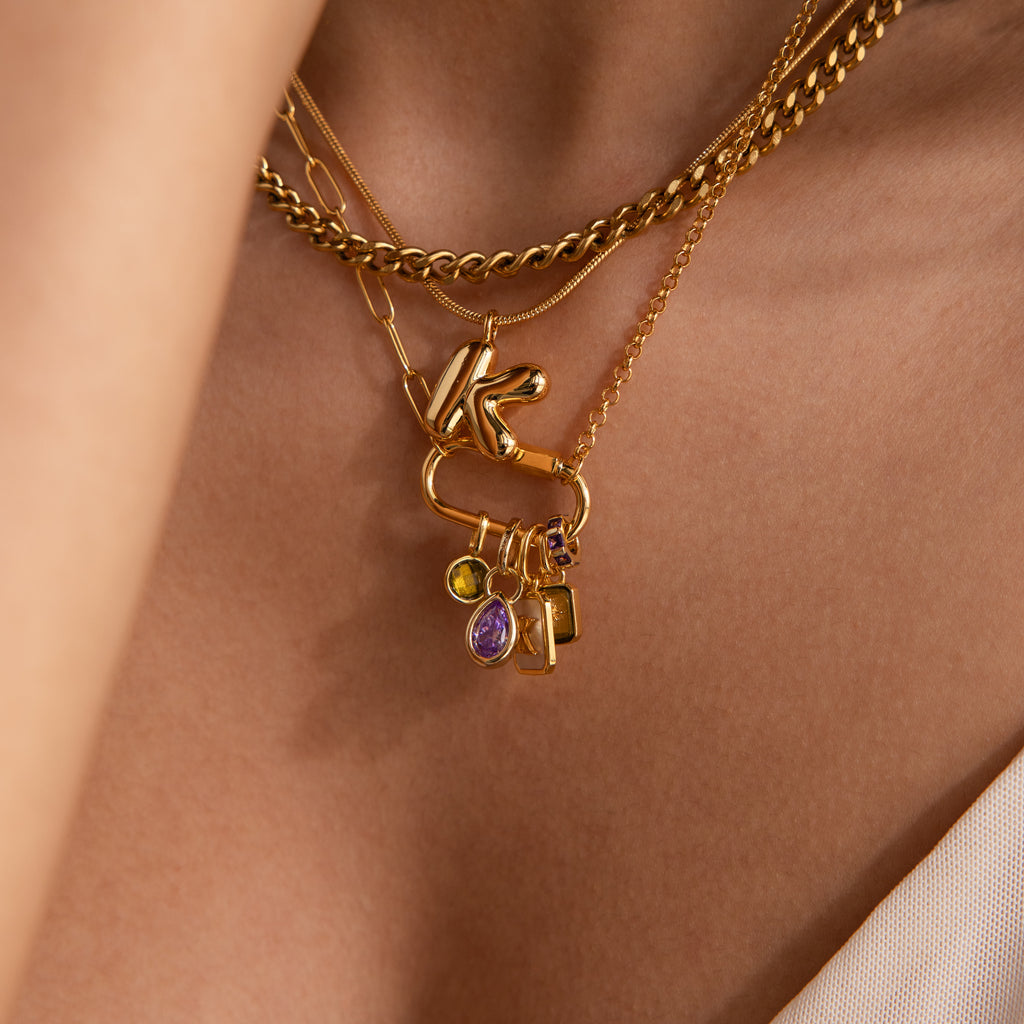 Close-up of layered gold necklaces with colorful charms, including the Infinity Princess Birthstone Charm, on a person’s neck wearing a light-colored top.