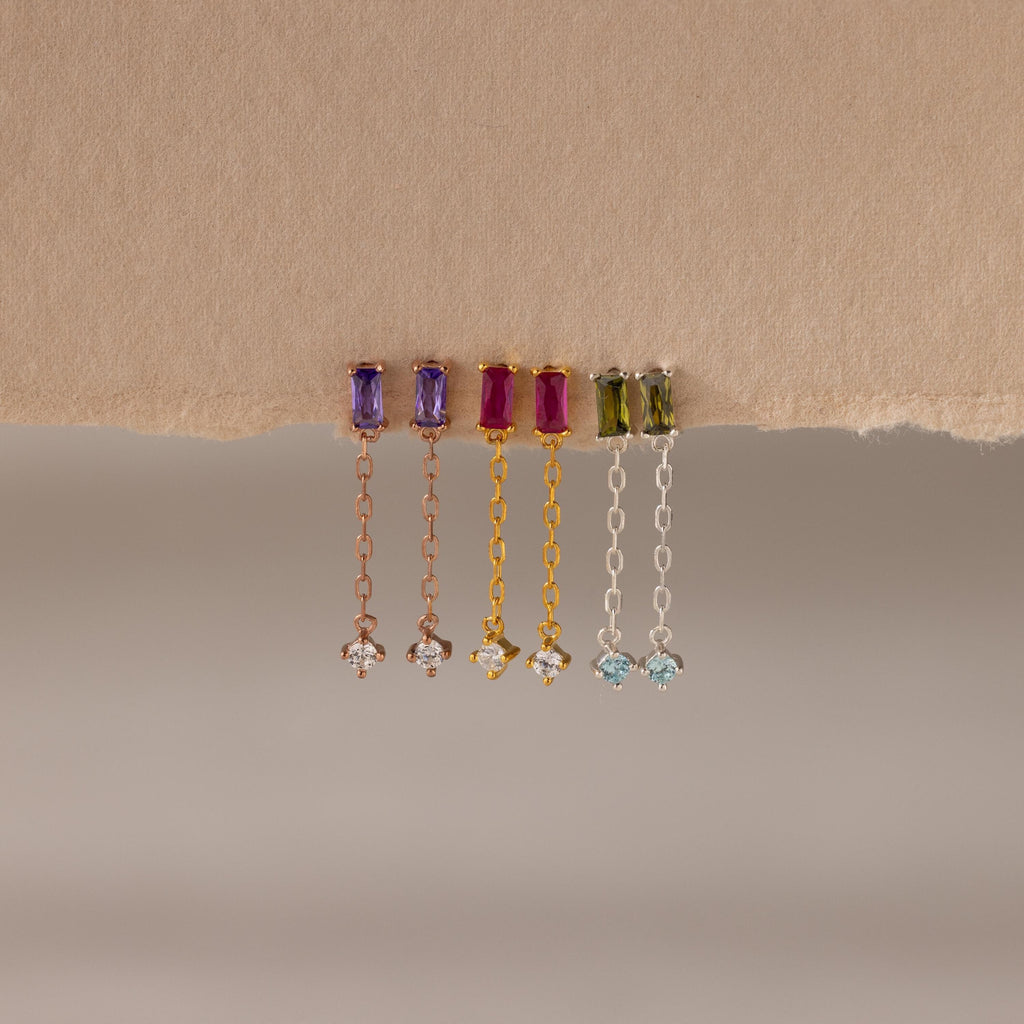 Four pairs of Duo Birthstone Drop Studs, each with vibrant baguette-cut gemstones and chain studs, hang elegantly from a torn piece of beige paper.