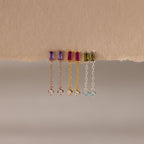 Four pairs of Duo Birthstone Drop Studs, each with vibrant baguette-cut gemstones and chain studs, hang elegantly from a torn piece of beige paper.