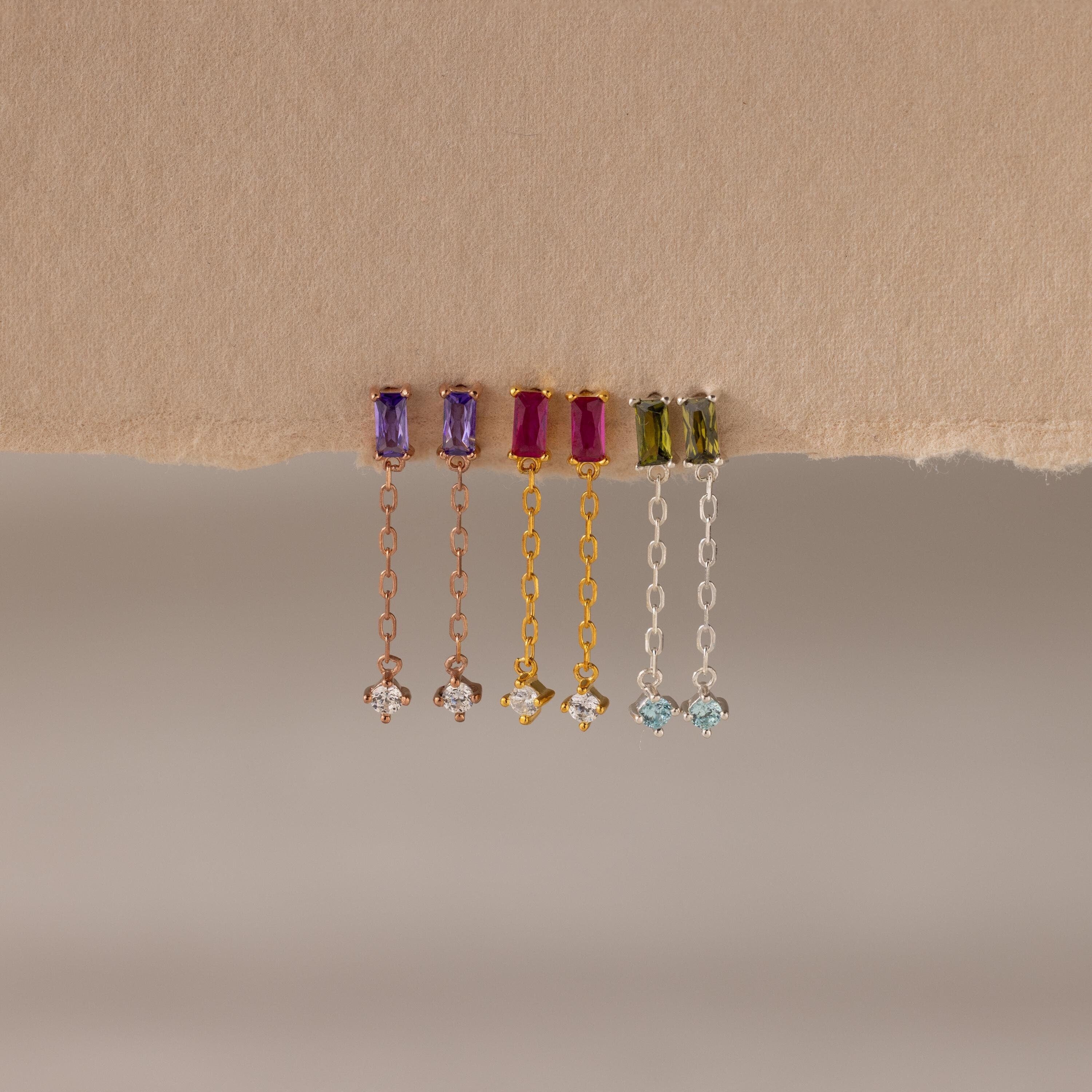 Four pairs of Duo Birthstone Drop Studs, each with vibrant baguette-cut gemstones and chain studs, hang elegantly from a torn piece of beige paper.