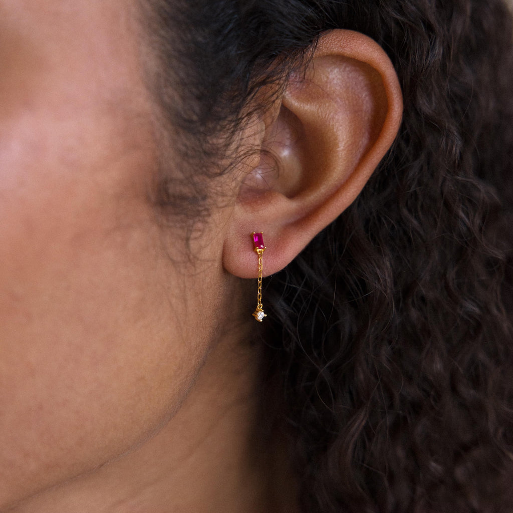 Close-up of an ear wearing Duo Birthstone Drop Studs: gold chain stud earrings with a small red gem and a dangling star charm.