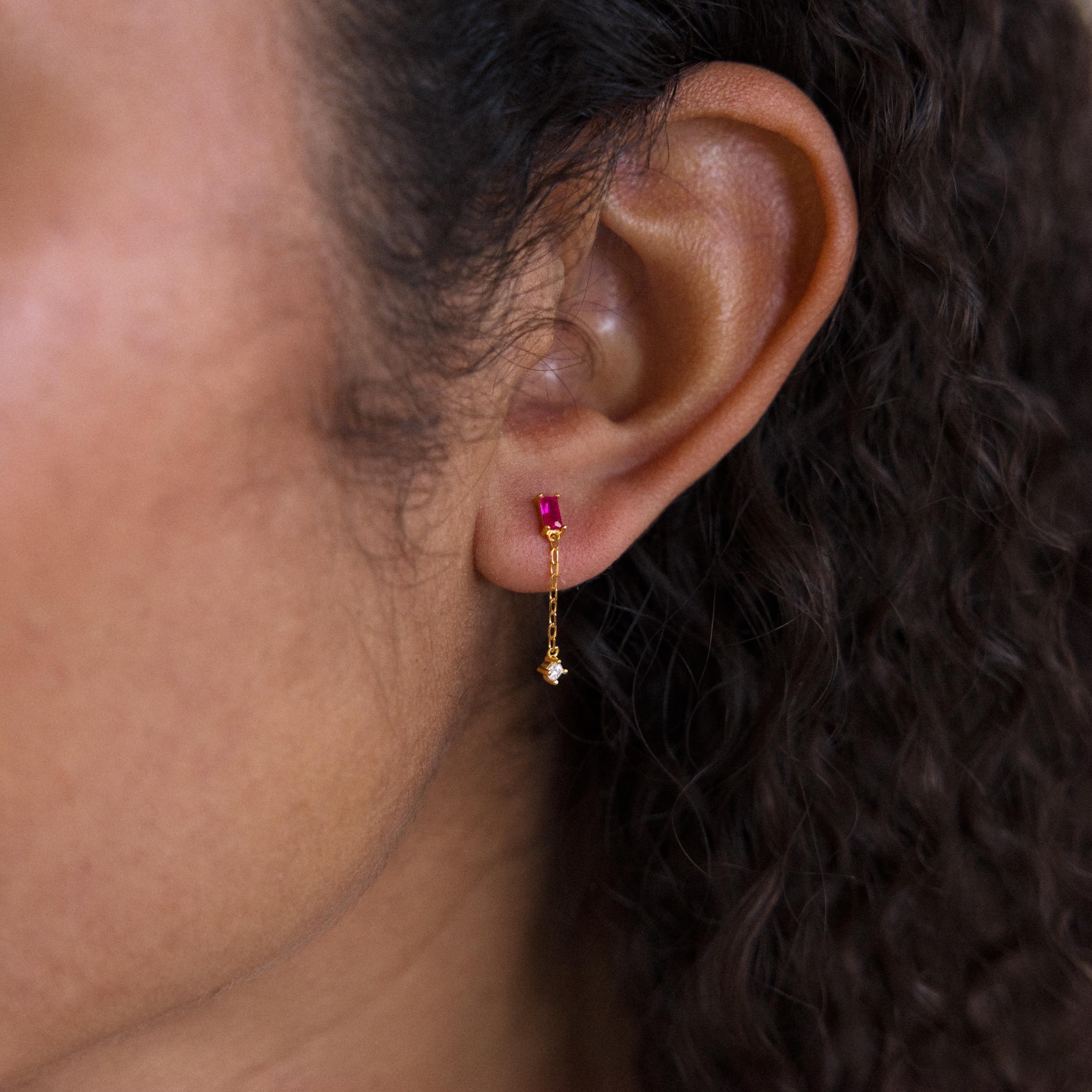 Close-up of an ear wearing Duo Birthstone Drop Studs: gold chain stud earrings with a small red gem and a dangling star charm.