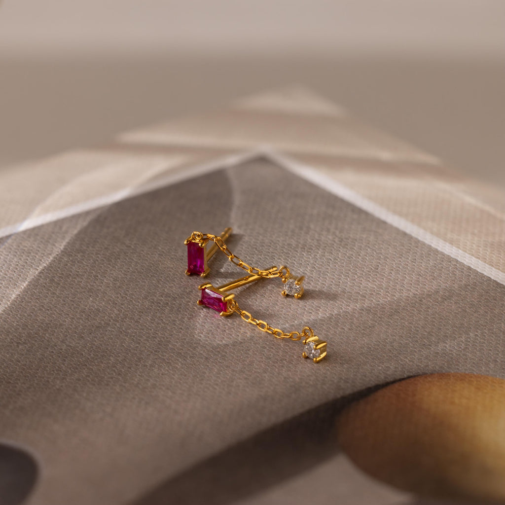 Duo Birthstone Drop Studs featuring gold chain, pink baguette-cut gemstones, and small diamonds, shown on a folded, blurred backdrop.