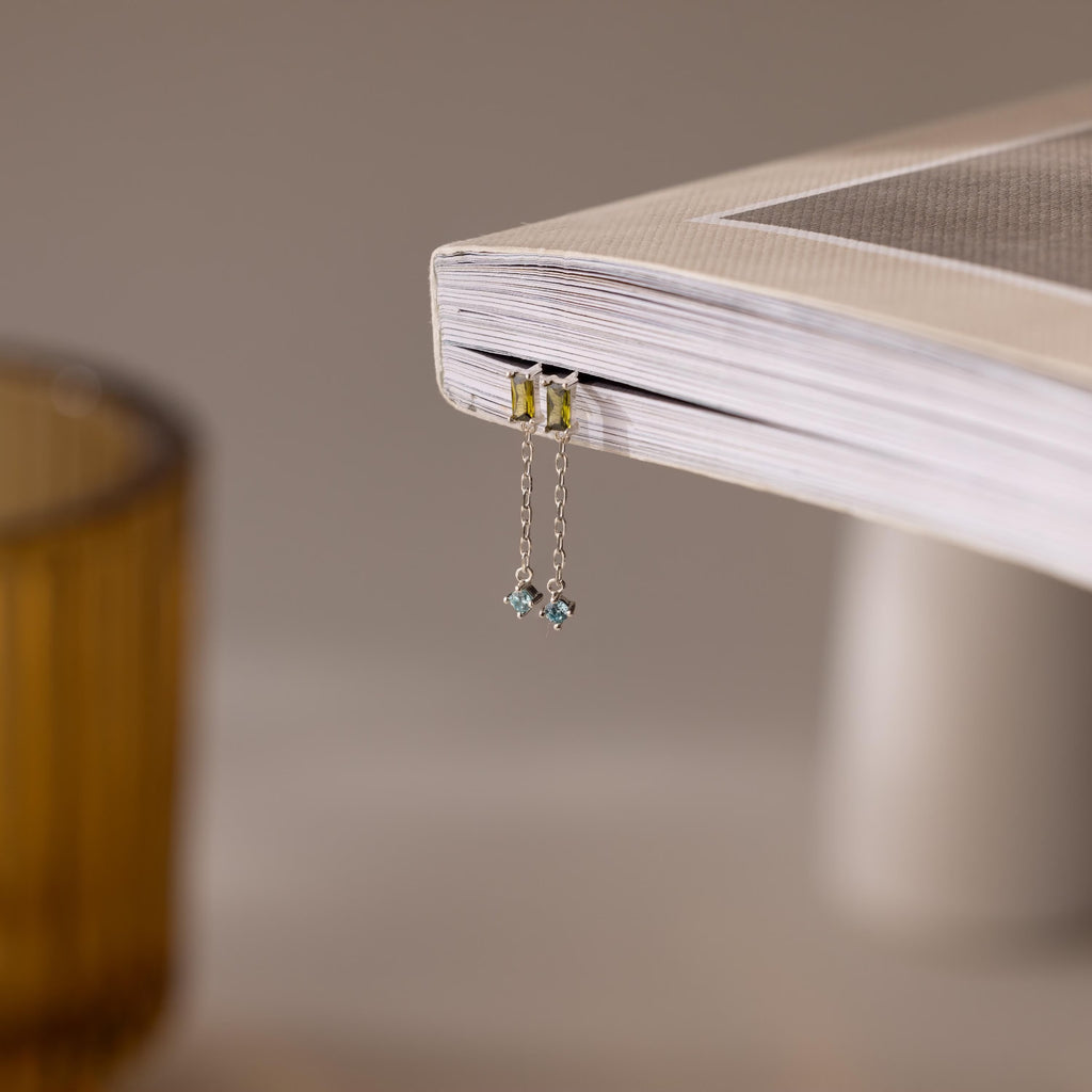The Duo Birthstone Drop Studs rest on the edge of a closed book, their delicate design and sparkling stones catching the light beautifully.