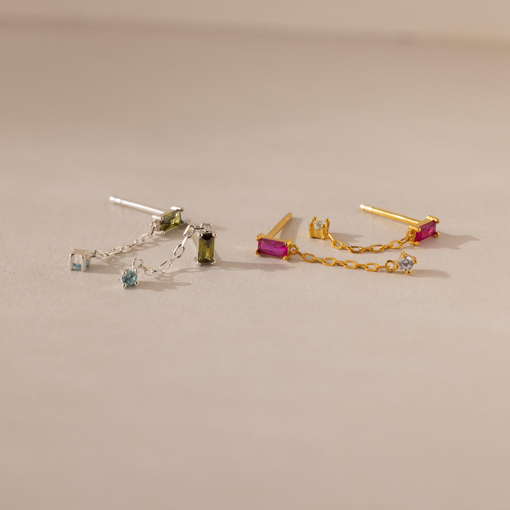 Duo Birthstone Drop Studs: one silver with blue and green baguette gems, one gold with pink and clear stones, displayed on a beige background.
