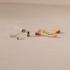 Duo Birthstone Drop Studs: one silver with blue and green baguette gems, one gold with pink and clear stones, displayed on a beige background.