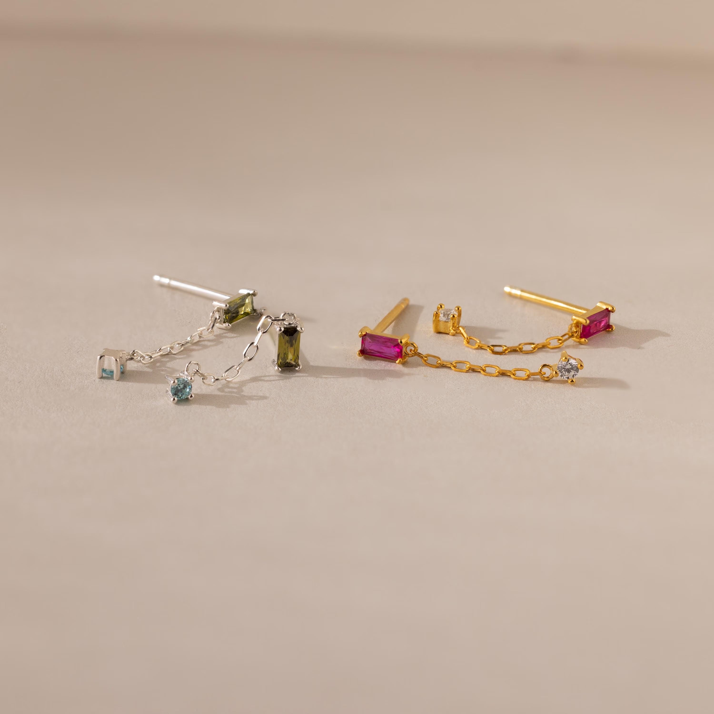 Duo Birthstone Drop Studs: one silver with blue and green baguette gems, one gold with pink and clear stones, displayed on a beige background.