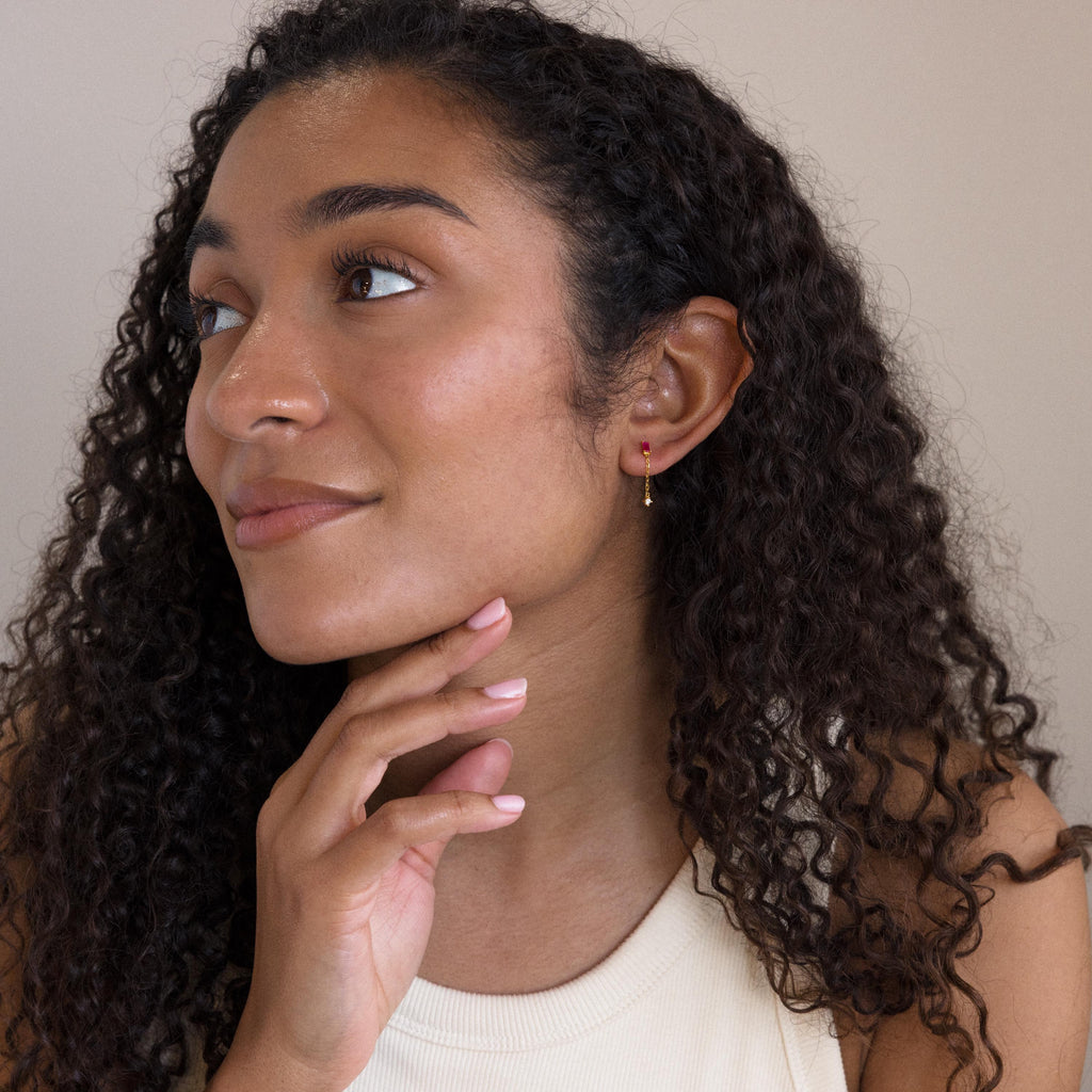 A woman with curly hair, smiling and looking to the side, wears a sleeveless top and the Duo Birthstone Drop Studs.